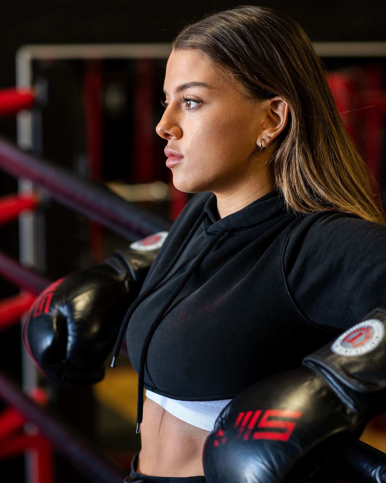 Female boxer in black crop hoodie wearing boxing gloves poses in gym near boxing ring.