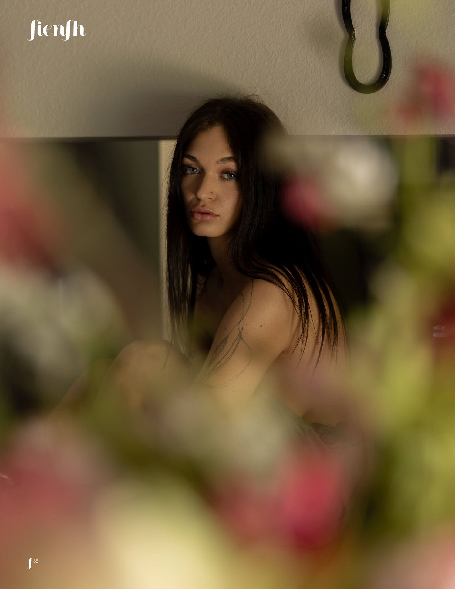 Young woman with long dark hair gazing thoughtfully, framed by soft blurred pink flowers in a moody artistic portrait.