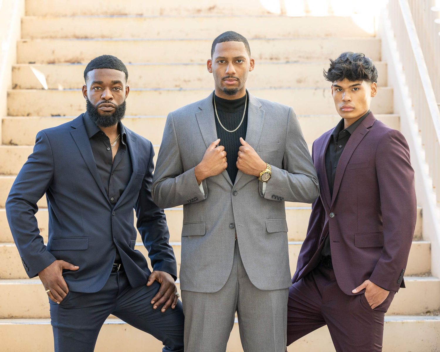 Three stylish men in suits posing on staircase in navy, gray, and burgundy outfits.