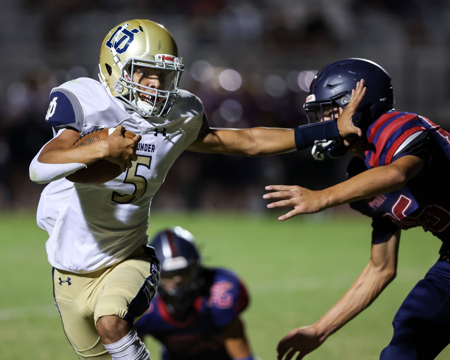 Football player #5 in gold uniform stiff-arms a defender during a night game.