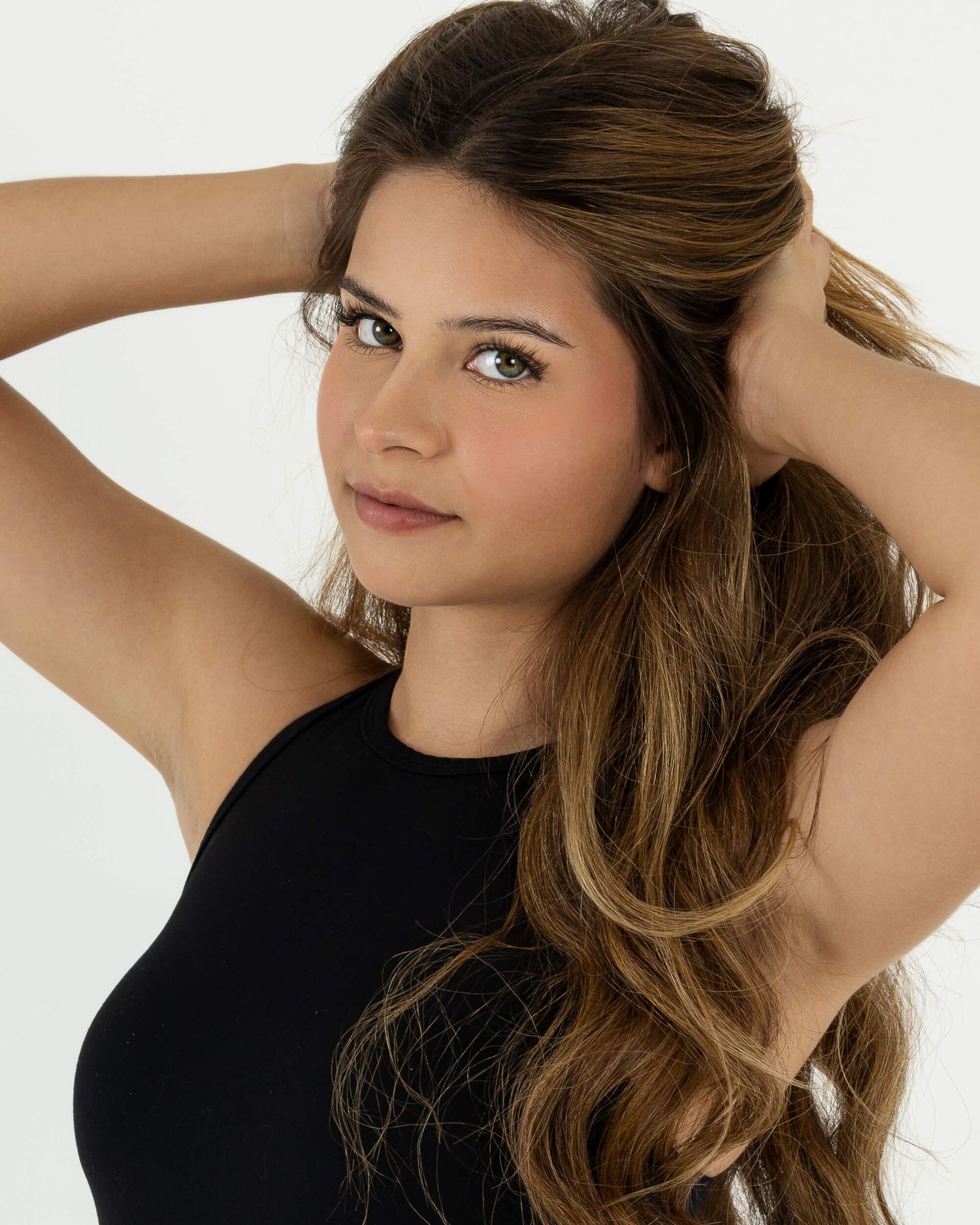 Young woman with long wavy brown hair in a black top poses with hands in hair for a modeling shot.