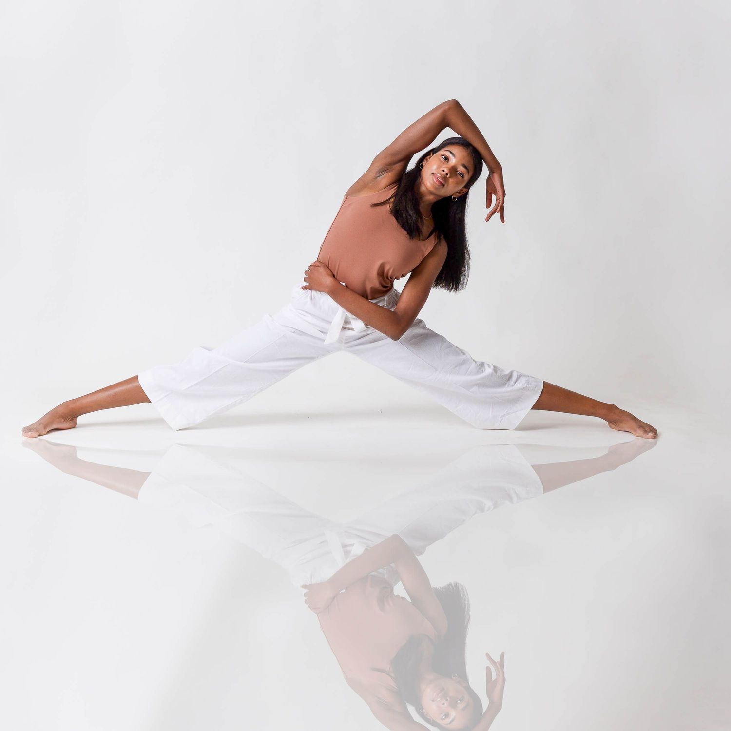 Flexible dancer performs a wide split stretch in brown top and white pants on a reflective white studio floor.