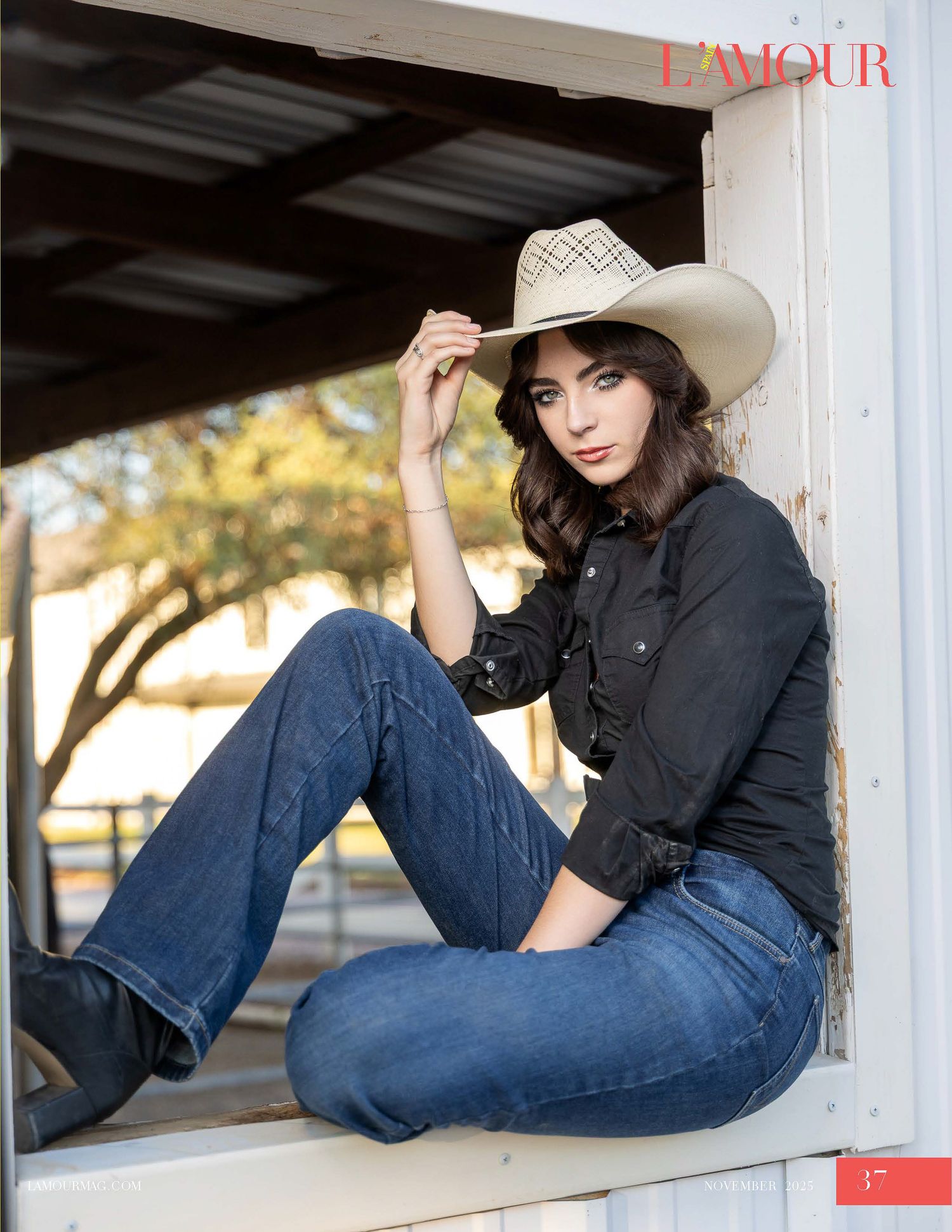 Brunette woman in black shirt, jeans, and cowboy hat poses in rustic barn for L'Amour magazine.