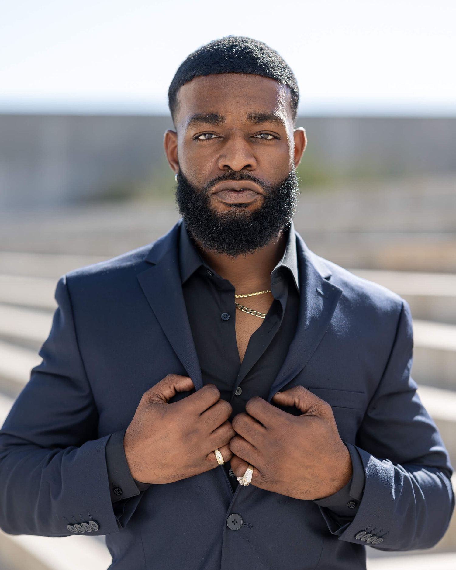 Man in navy suit adjusting jacket lapels outdoors, wearing gold chain necklace and ring.