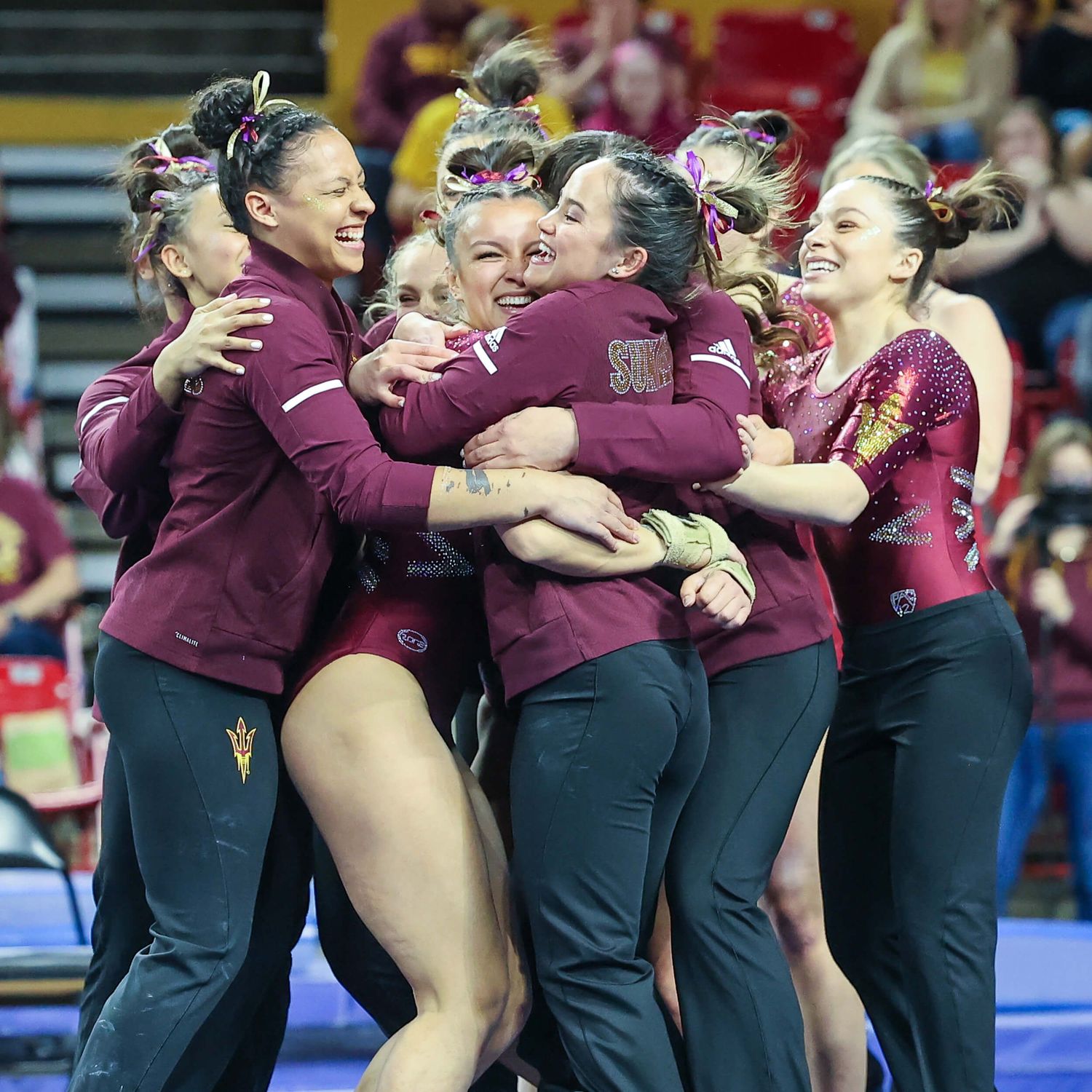 ASU gymnastics team celebrates together in a group hug wearing maroon warm-up jackets after a meet.