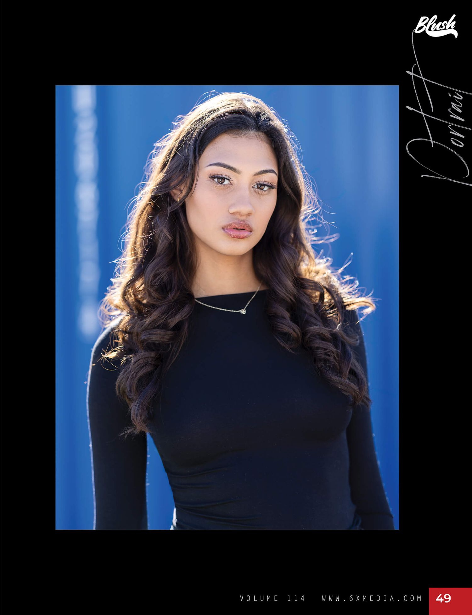 Young woman in black long-sleeve top poses against blue wall for Blesh portrait feature.