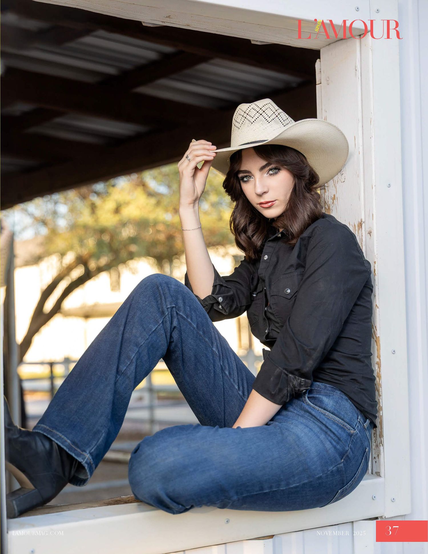 Brunette woman in black shirt, jeans, and cowboy hat sits on fence for L'Amour Magazine shoot.