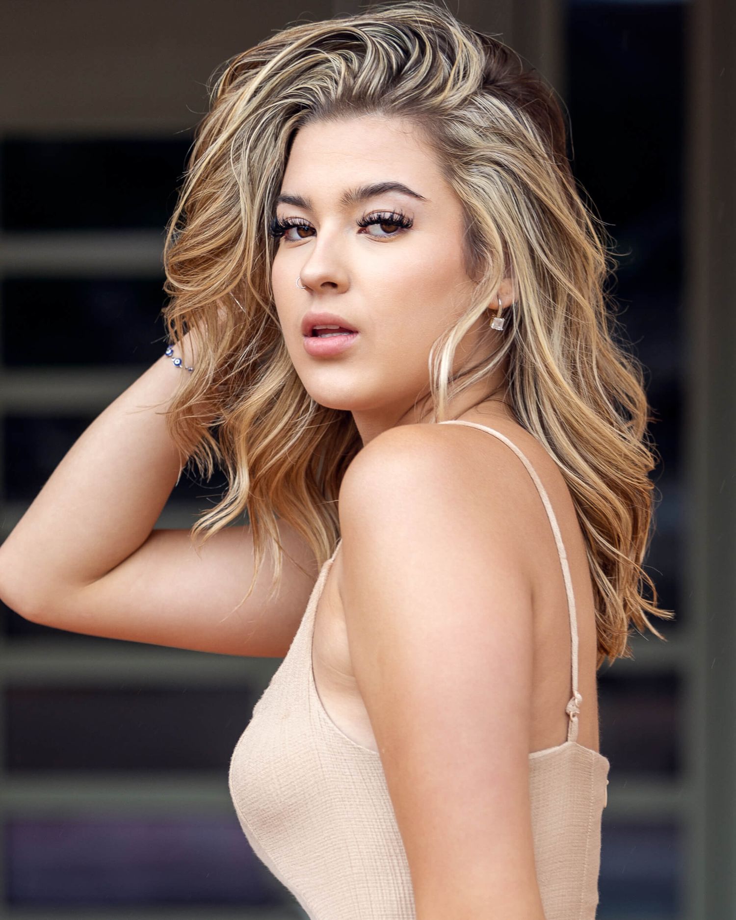 Young woman with highlighted wavy hair poses in a beige camisole with hand in hair.
