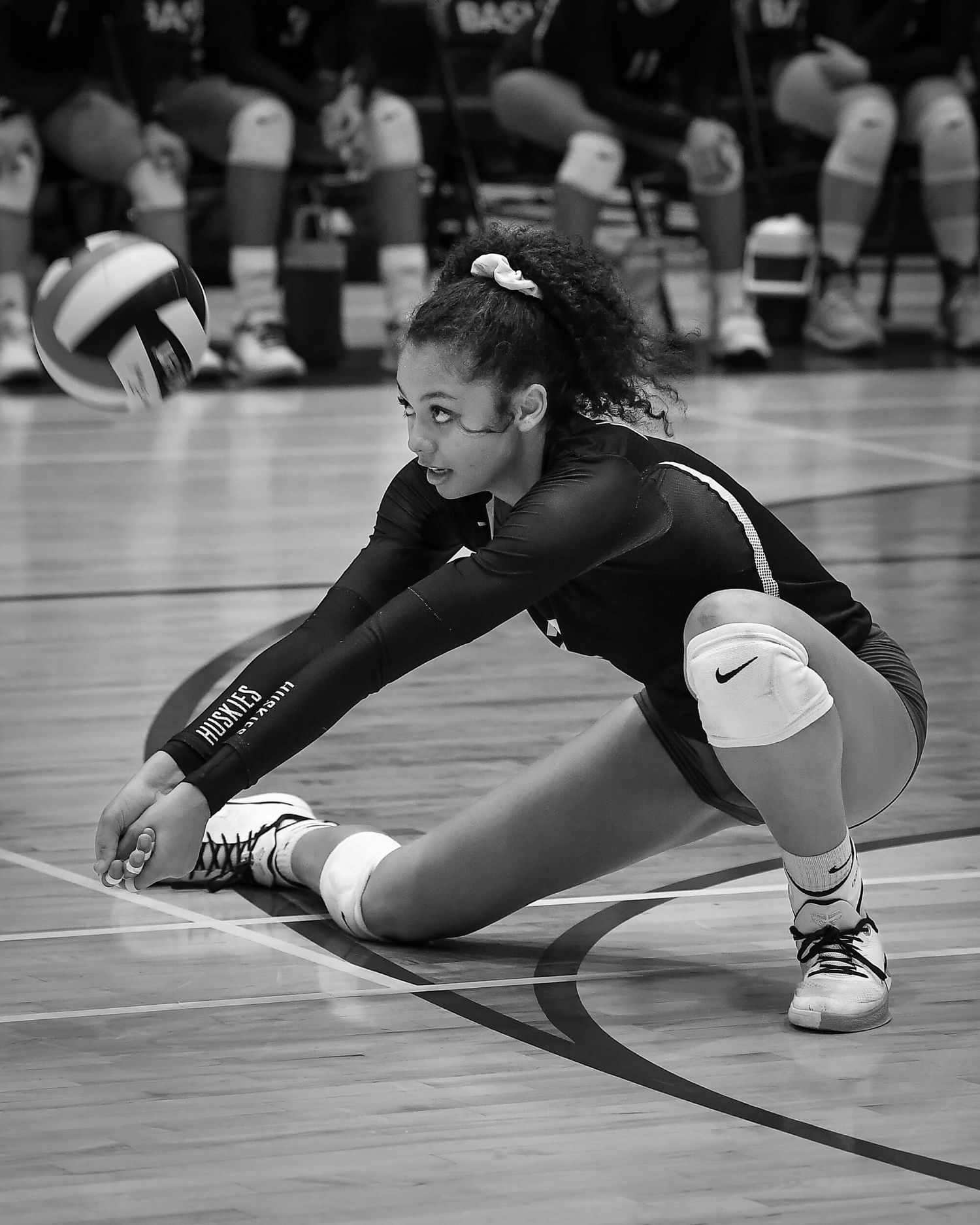 A female volleyball player digs low to receive the ball during an indoor match in black and white.