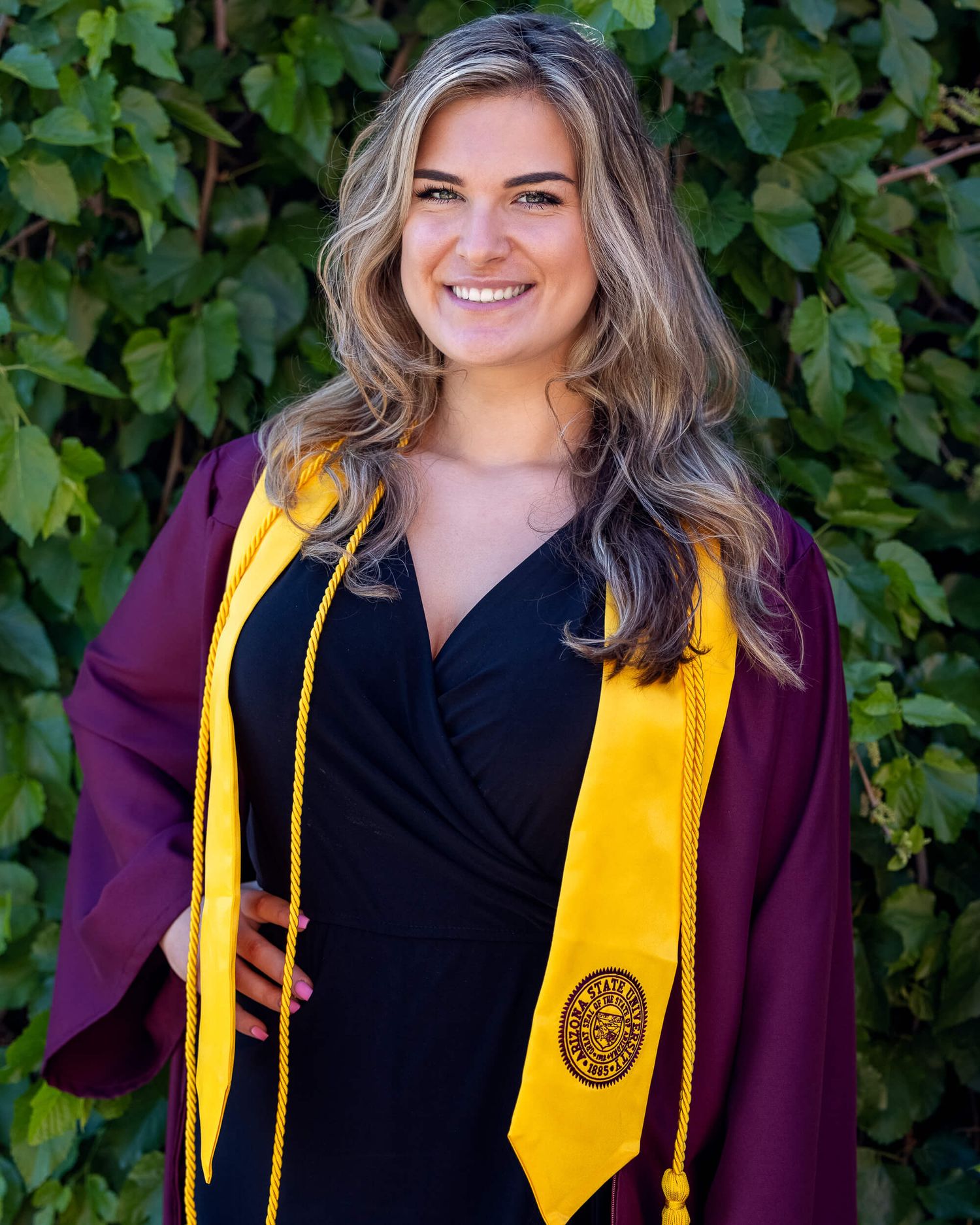 Woman in maroon graduation gown and gold stole smiling outdoors.
