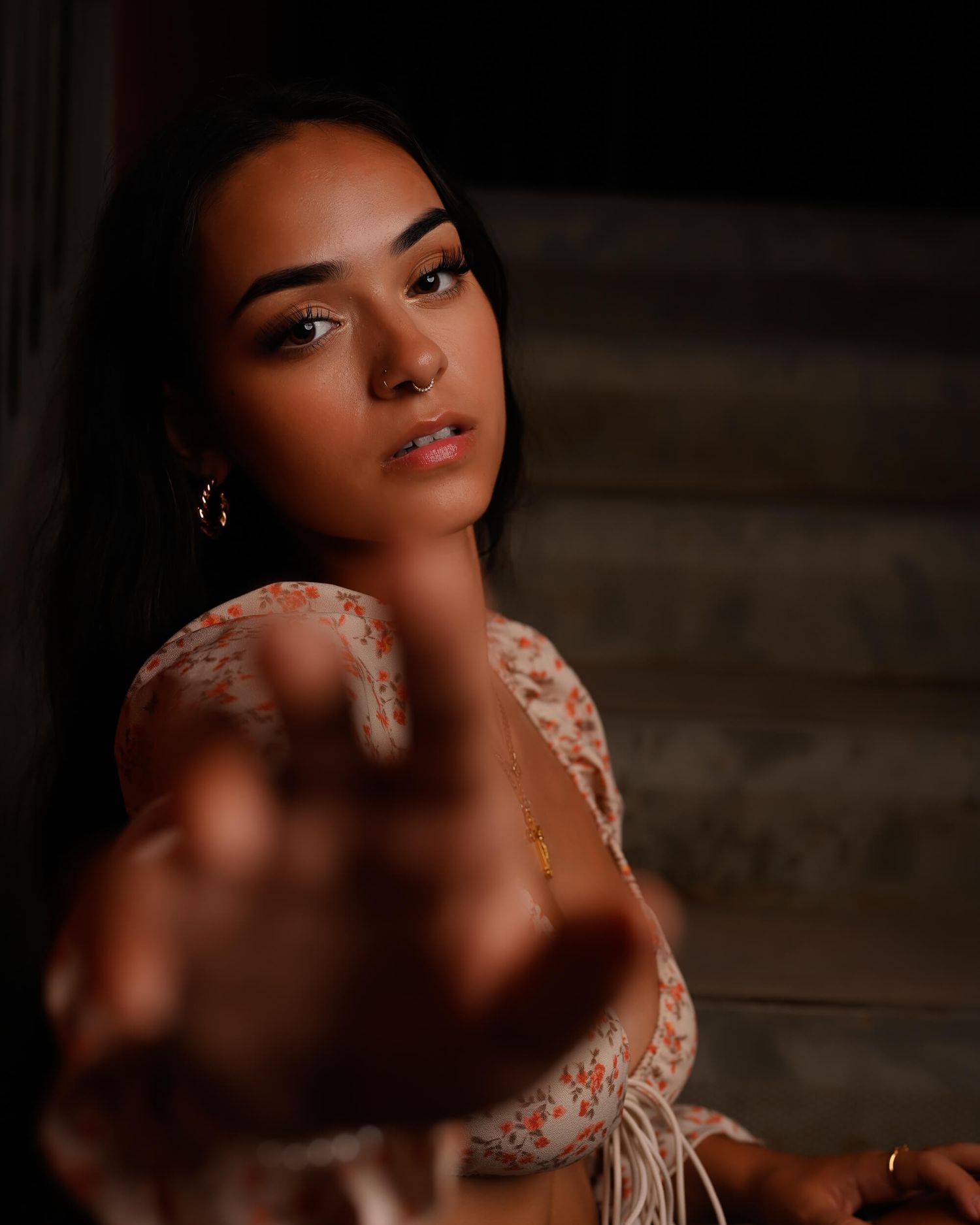 Young woman in floral top reaching toward camera on dark staircase.