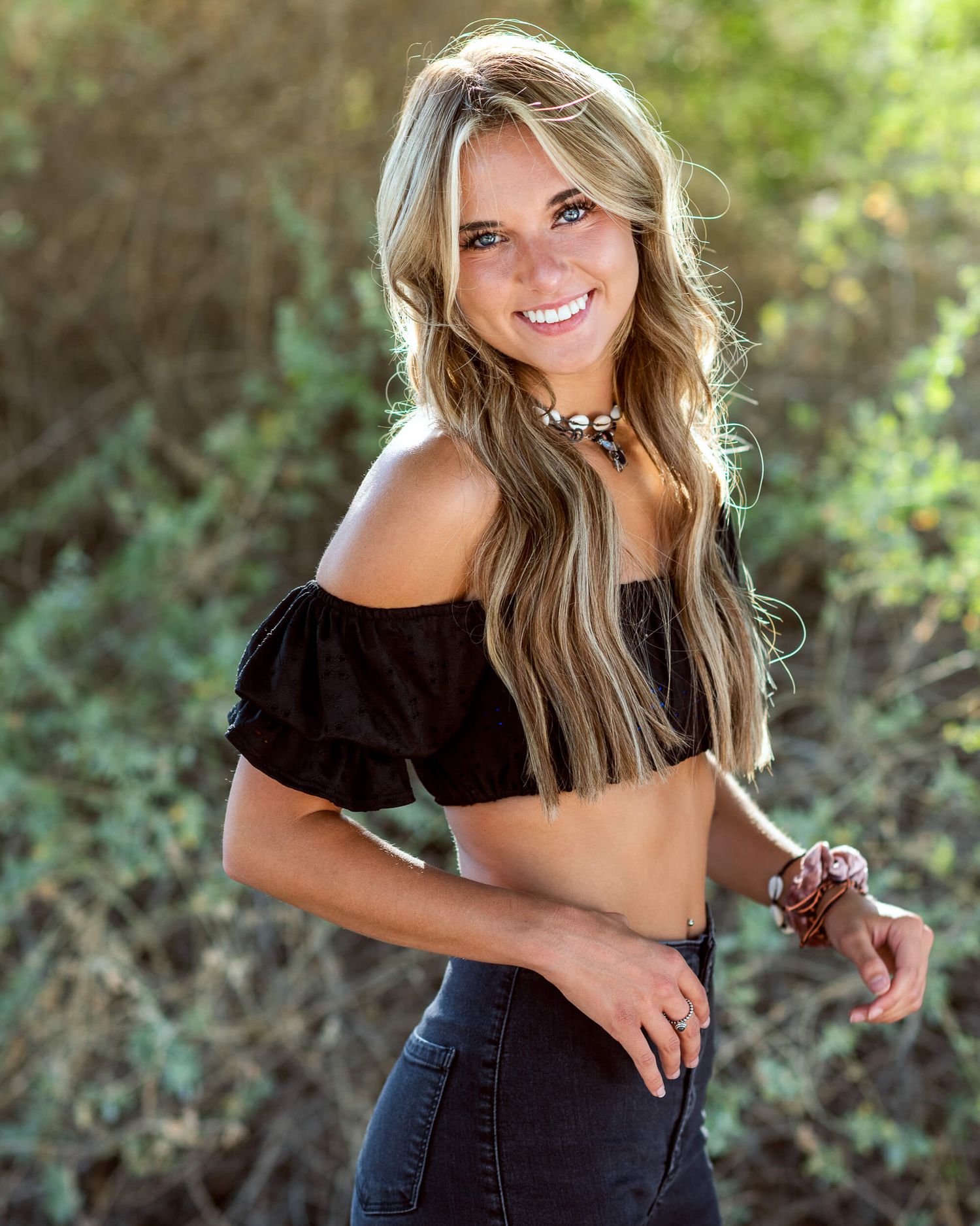 Smiling blonde woman in black off-shoulder crop top and jeans posing outdoors.