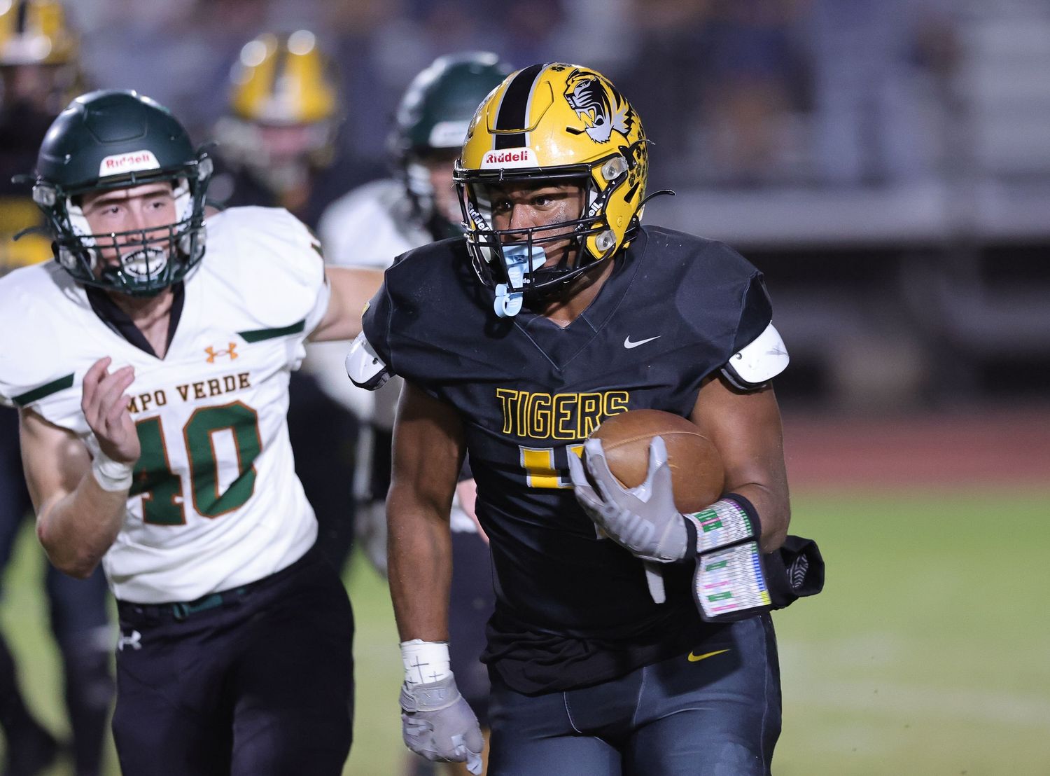 High school football player in black and gold Tigers uniform runs past a defender during a night game.