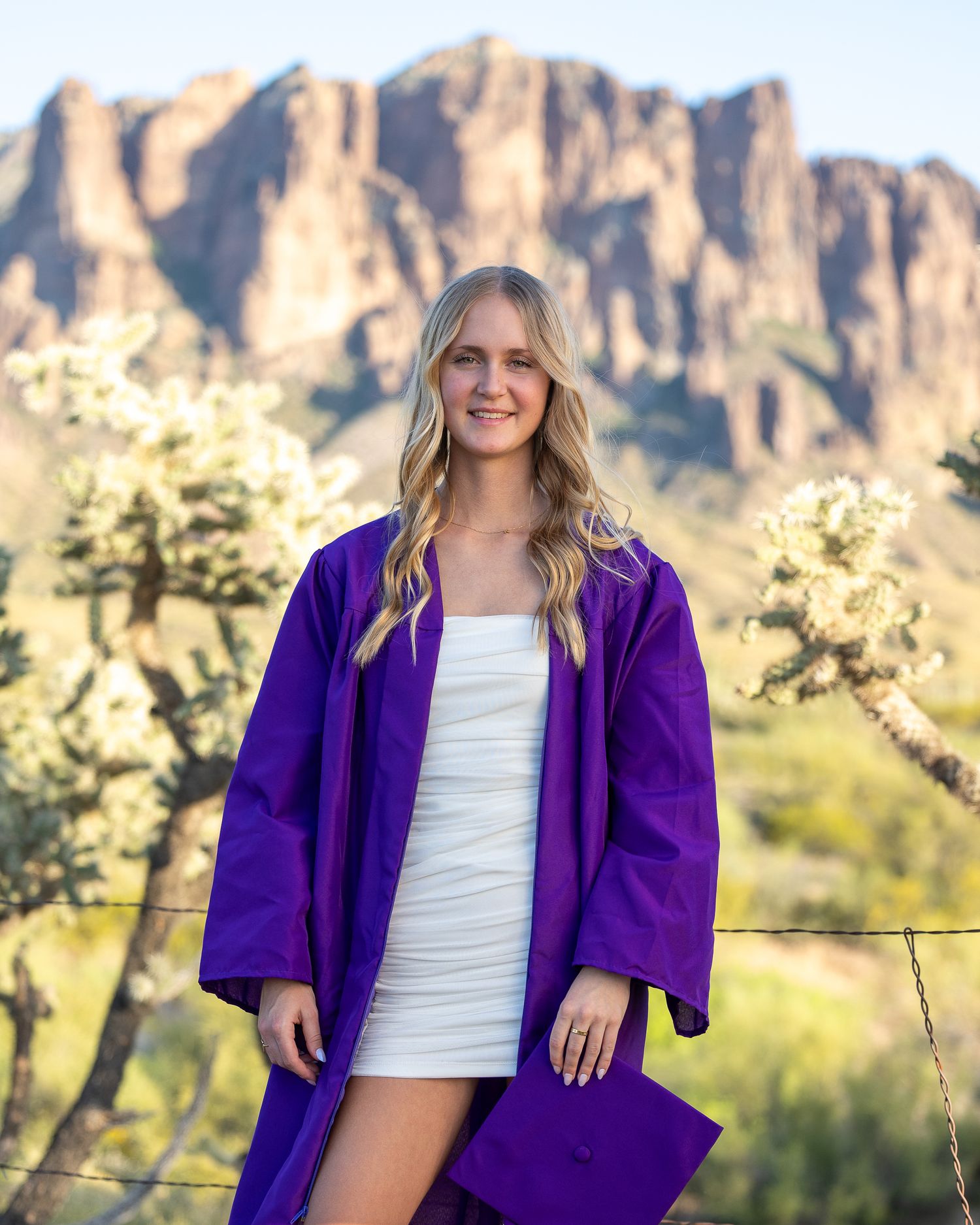 Smiling blonde woman in purple graduation gown posing in front of rocky mountain desert landscape.