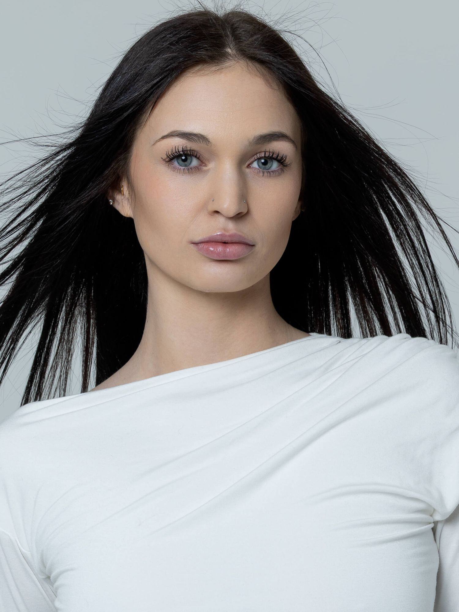 Young woman with long dark hair and blue eyes wearing a white top against a light gray background.