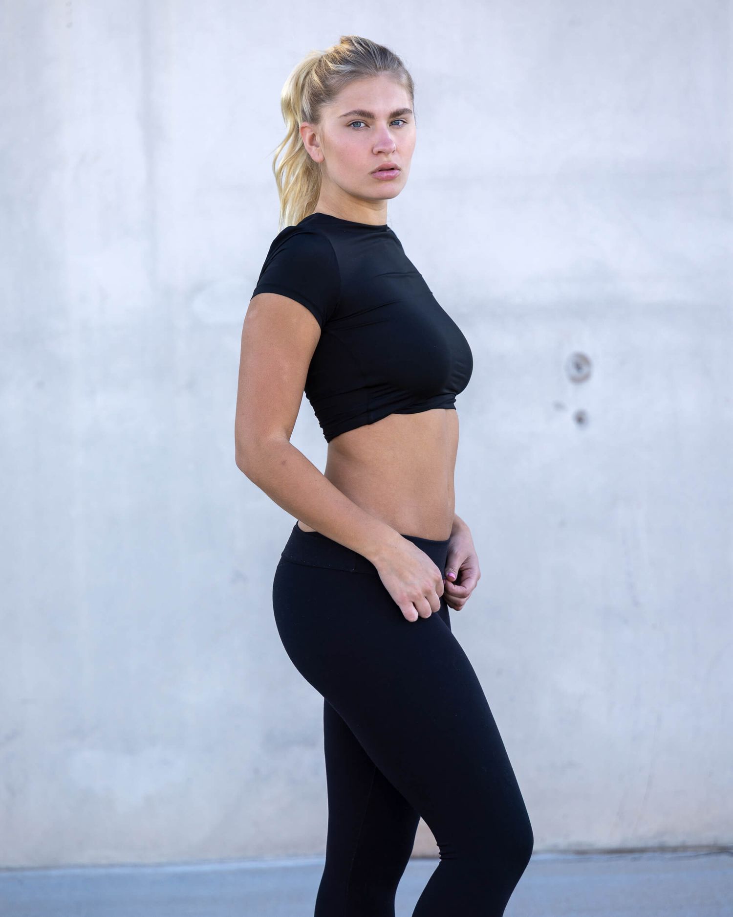 Woman in black crop top and leggings posing outdoors against a concrete wall in athletic wear.