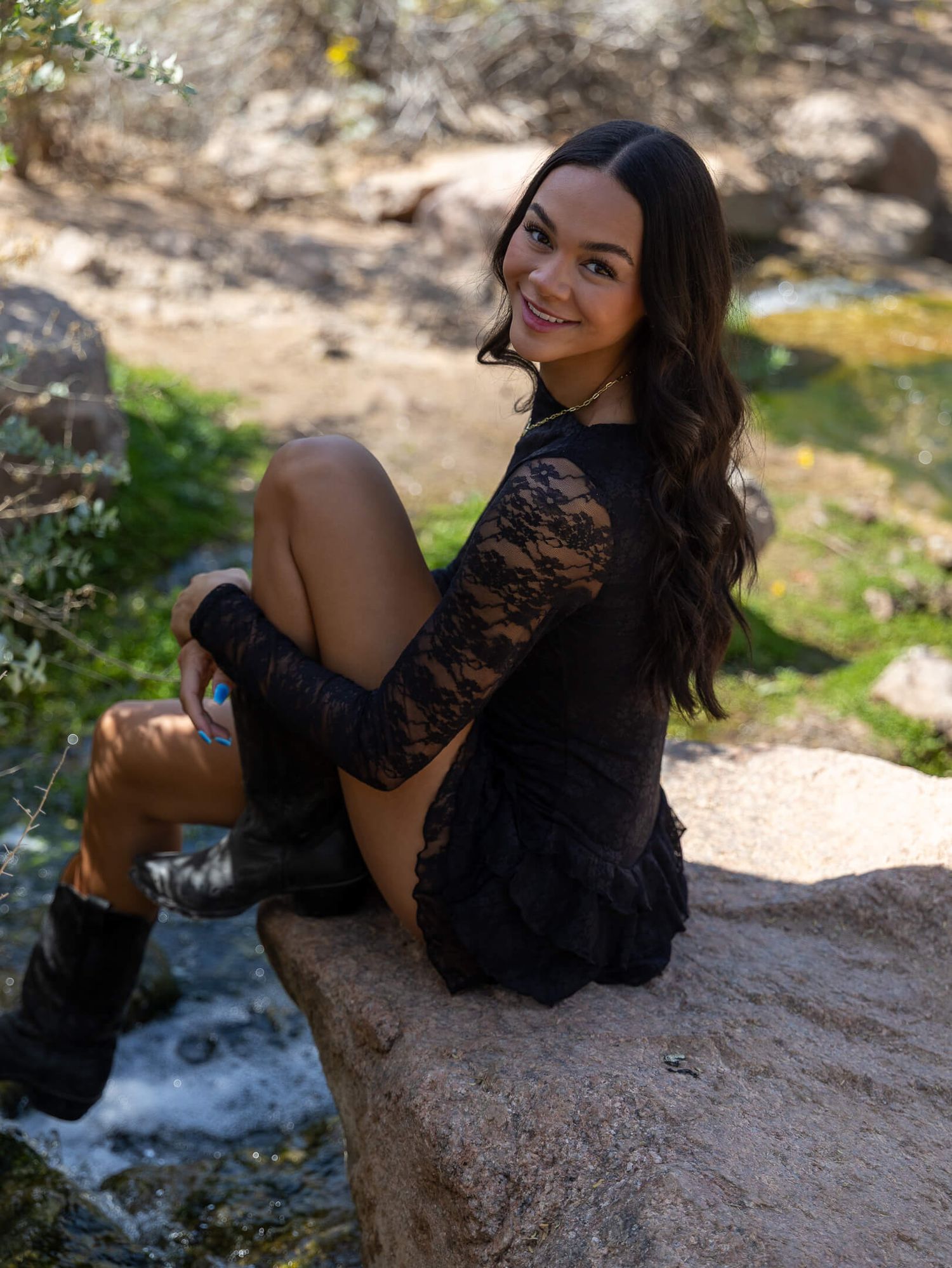Young woman in black lace dress and boots sits on rock near stream in nature setting.