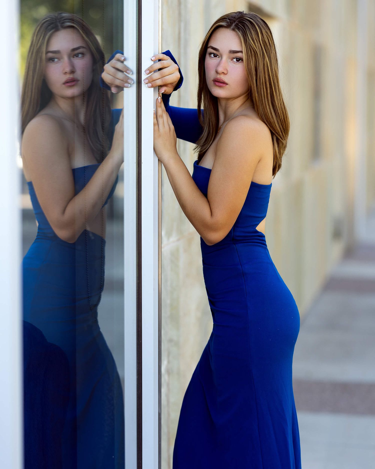 Elegant woman in blue strapless gown posing beside a reflective glass surface outdoors.