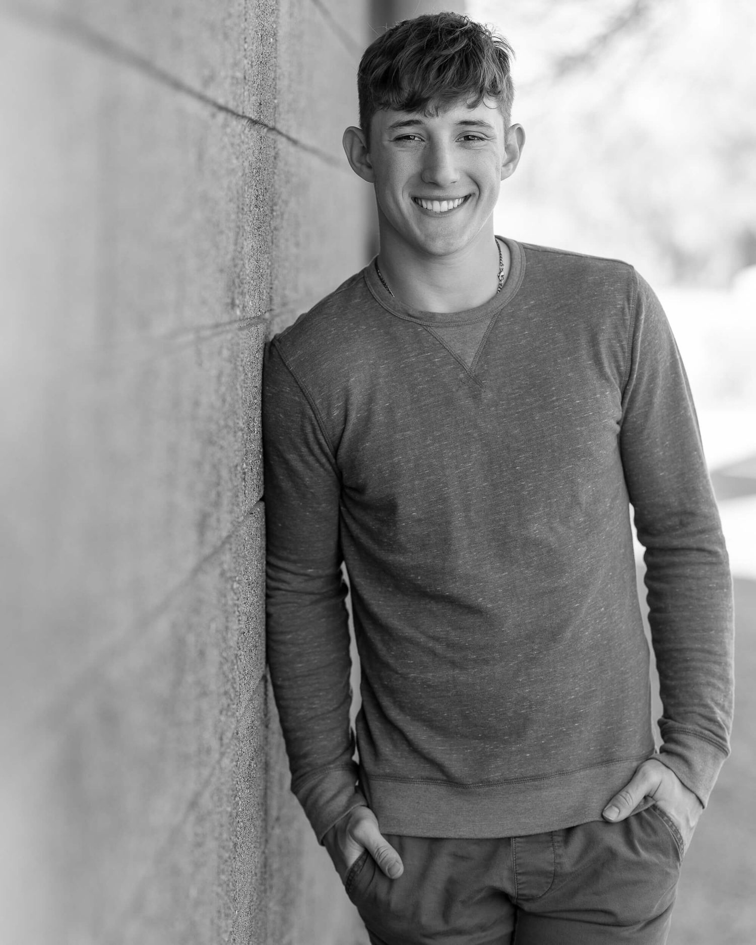 Smiling teen boy black and white senior photo leaning against wall in gray sweatshirt.