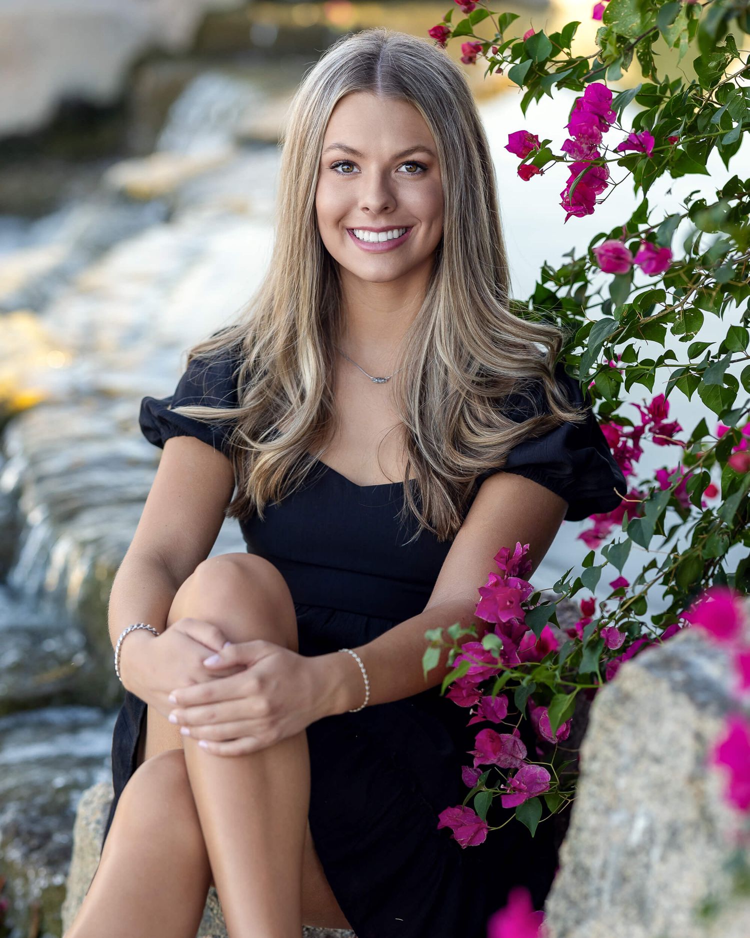 Young woman in black dress sitting on rocks near waterfall surrounded by pink bougainvillea flowers.