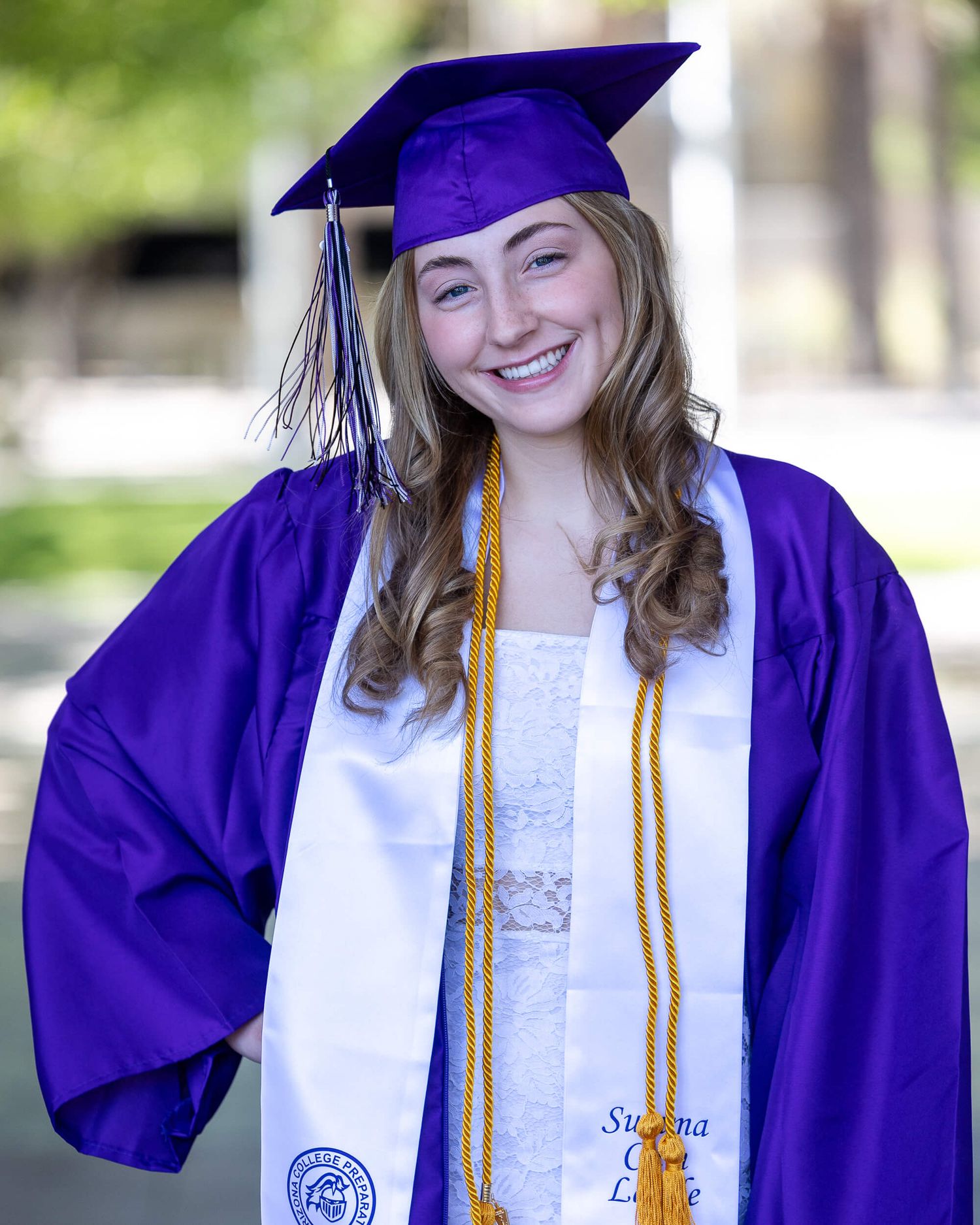 Smiling young woman in purple graduation cap and gown with gold honor cords and white stole.