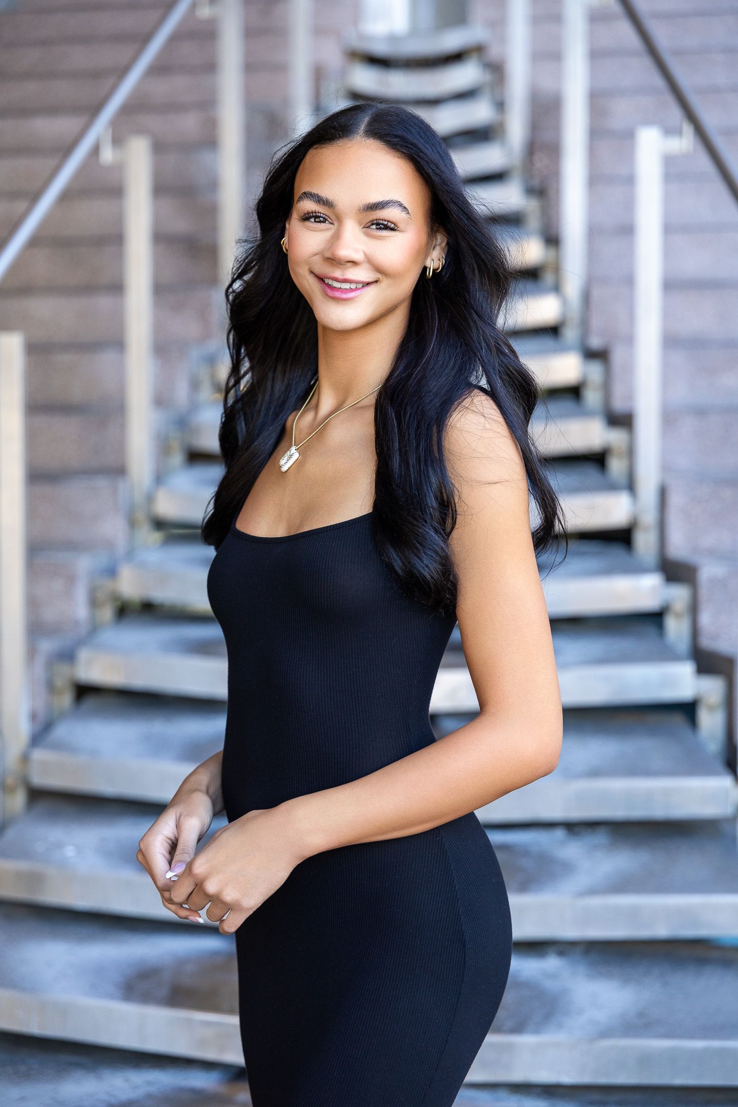 Young woman in black dress smiling outdoors near wooden staircase, elegant portrait photography.