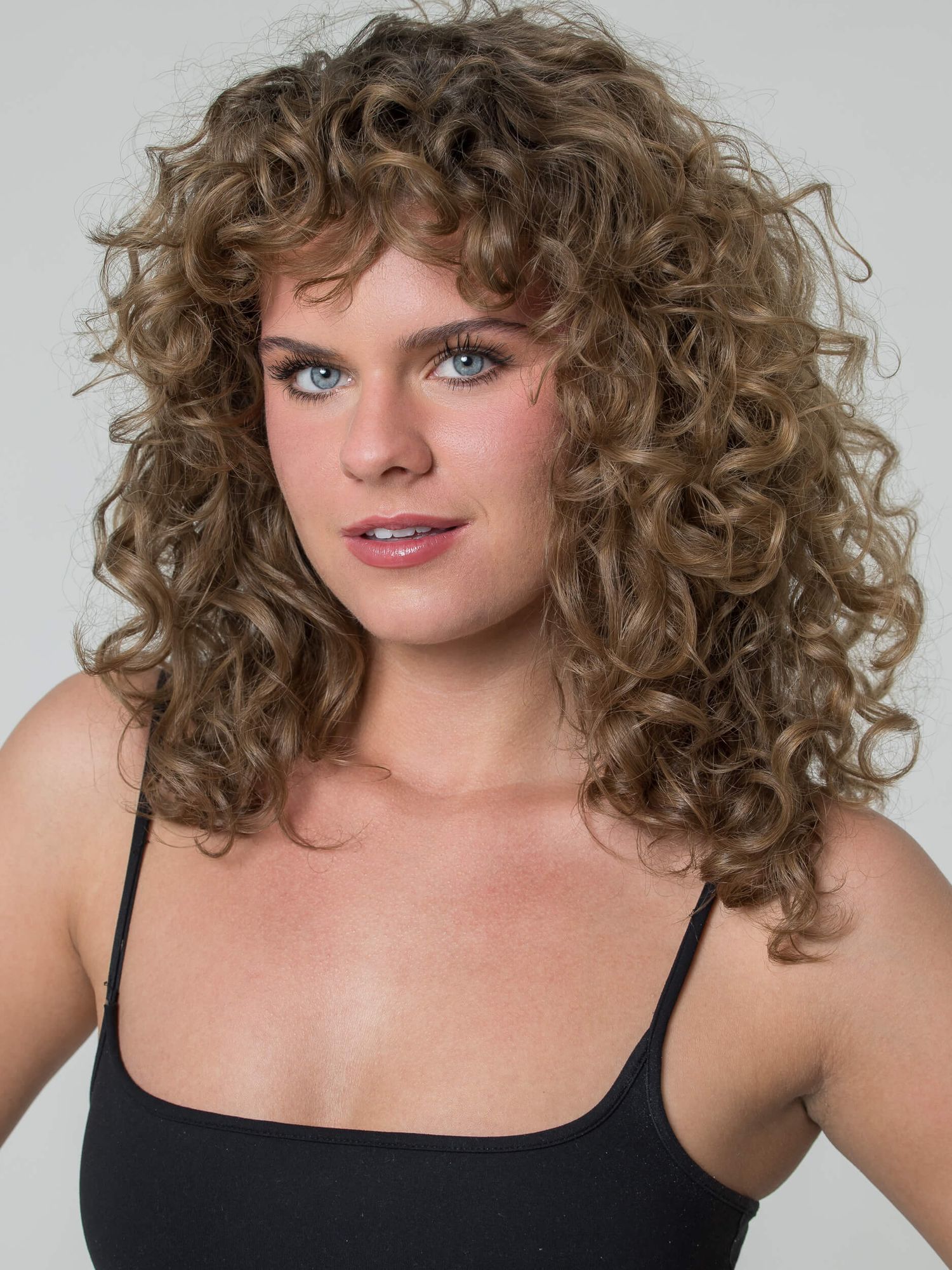 Woman with voluminous curly brown hair and bangs wearing a black top, showcasing a natural curly hairstyle.