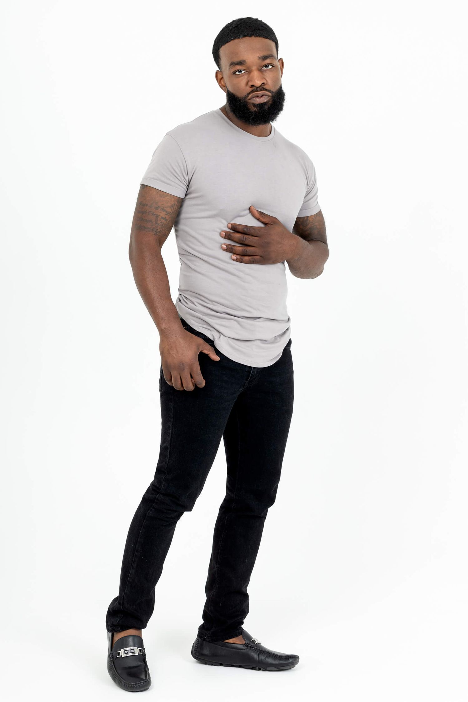 Bearded Black man in gray t-shirt and dark jeans poses confidently against white background.