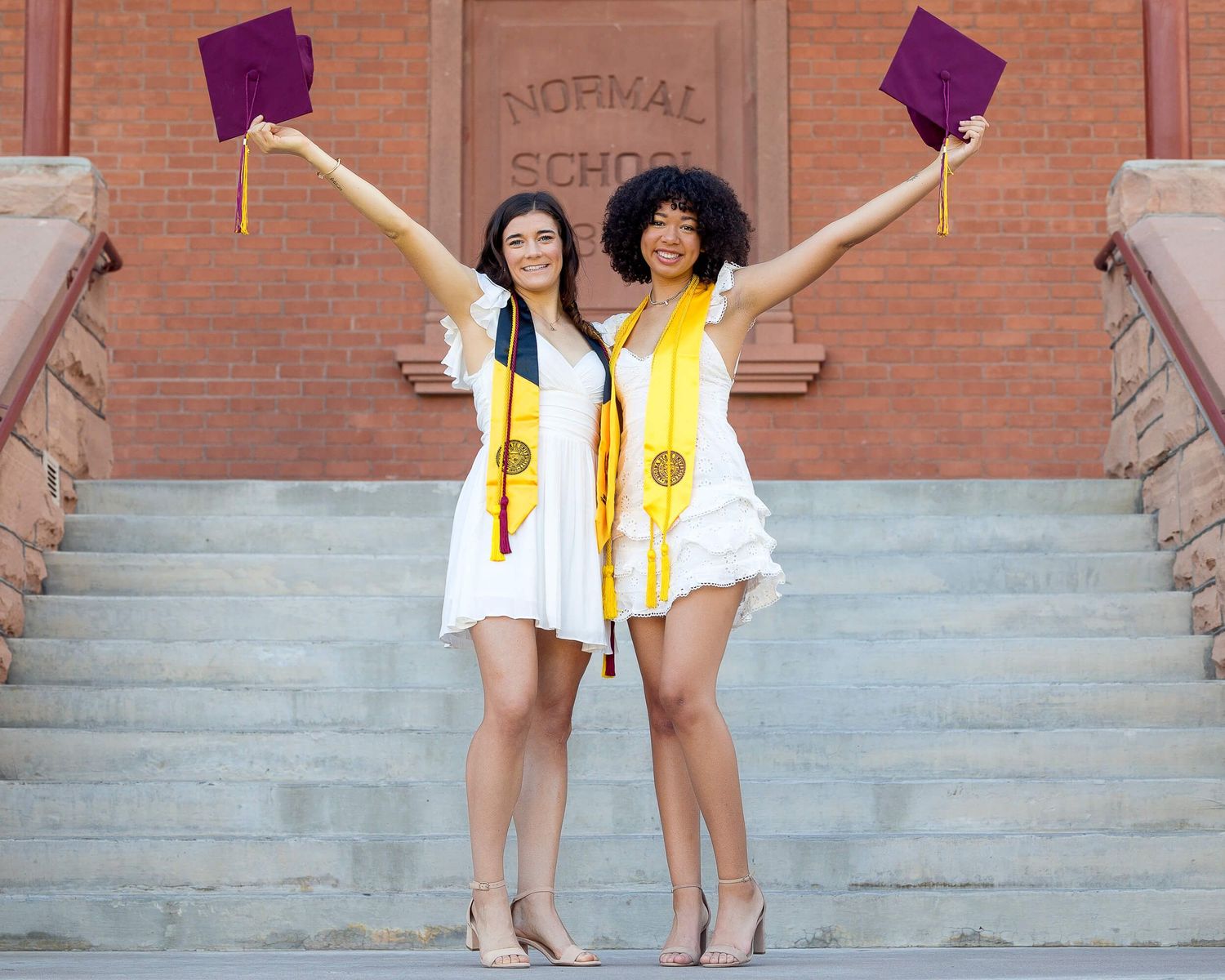 Two smiling graduates in white dresses raising maroon caps in front of Normal School historic building.