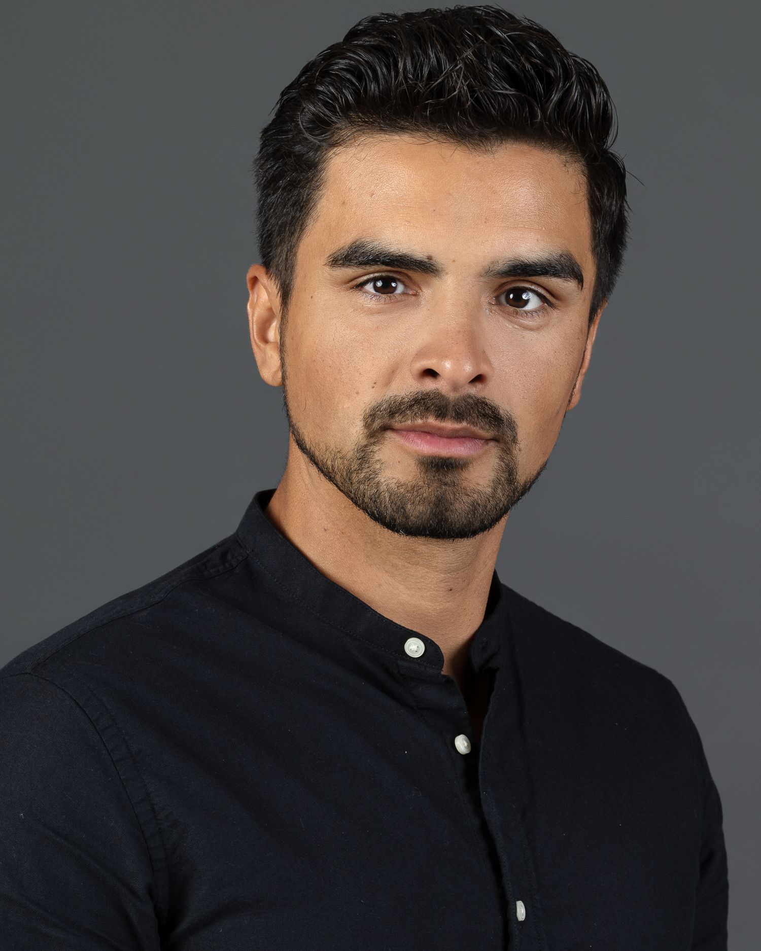 Professional headshot of young man with beard wearing black button-up shirt on gray background.