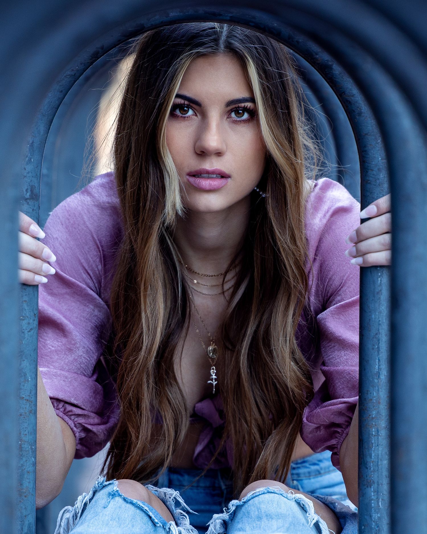 Brunette woman in purple blouse and ripped jeans posing in dramatic arched metal doorway.