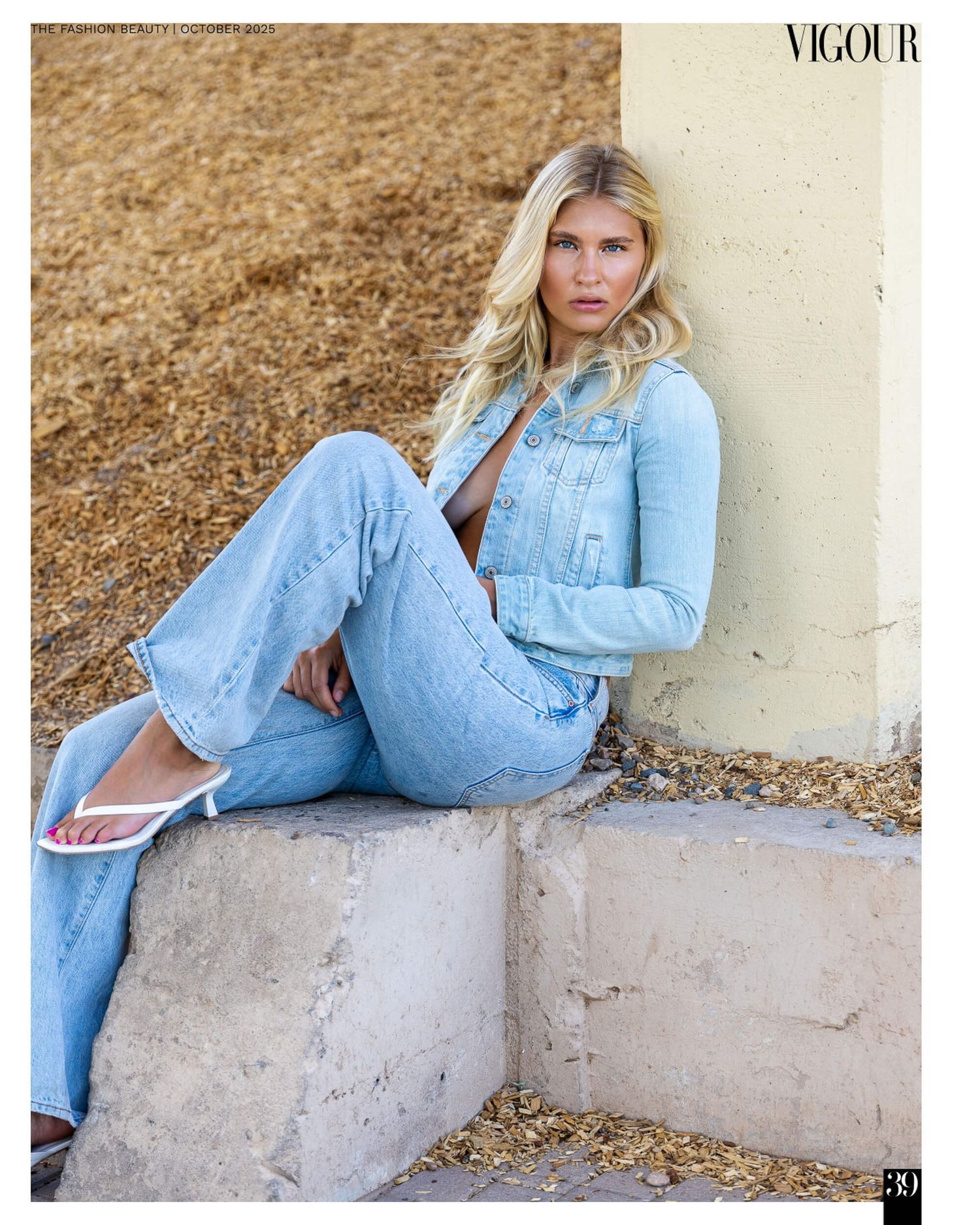 Blonde model in light wash denim jacket and flared jeans poses against a wall in Vigour magazine.