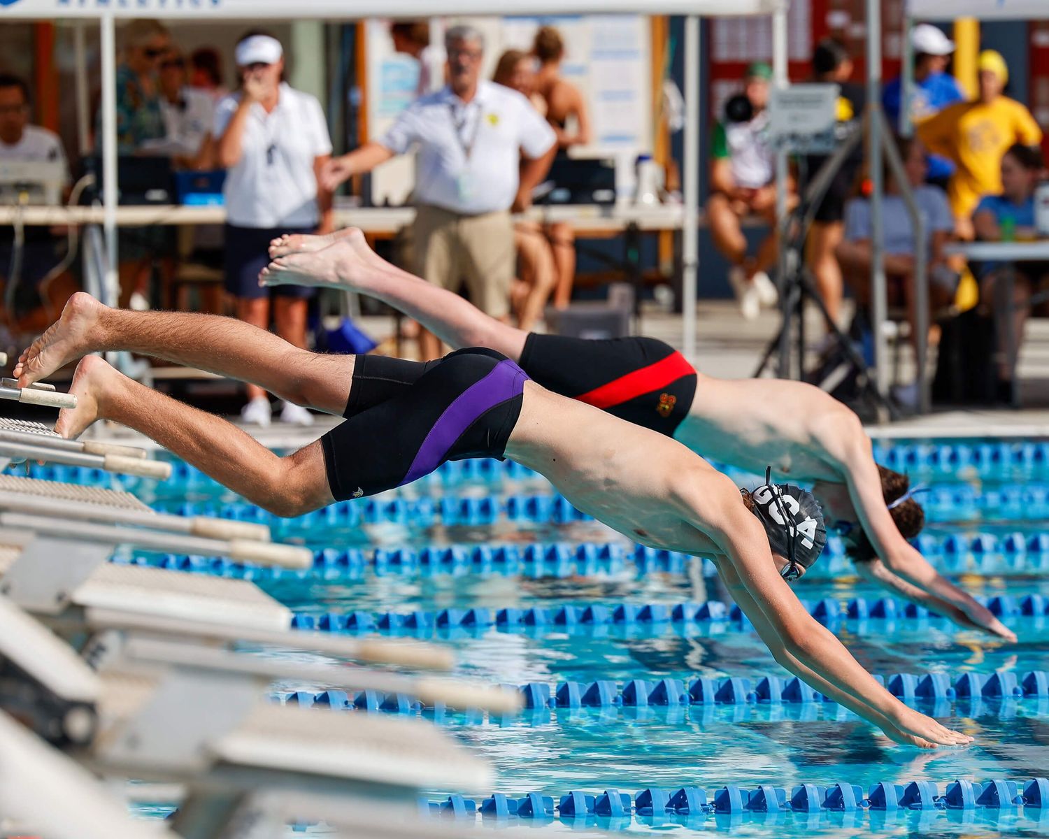 Competitive swimmers dive into a pool at the start of a race during an outdoor swim meet.