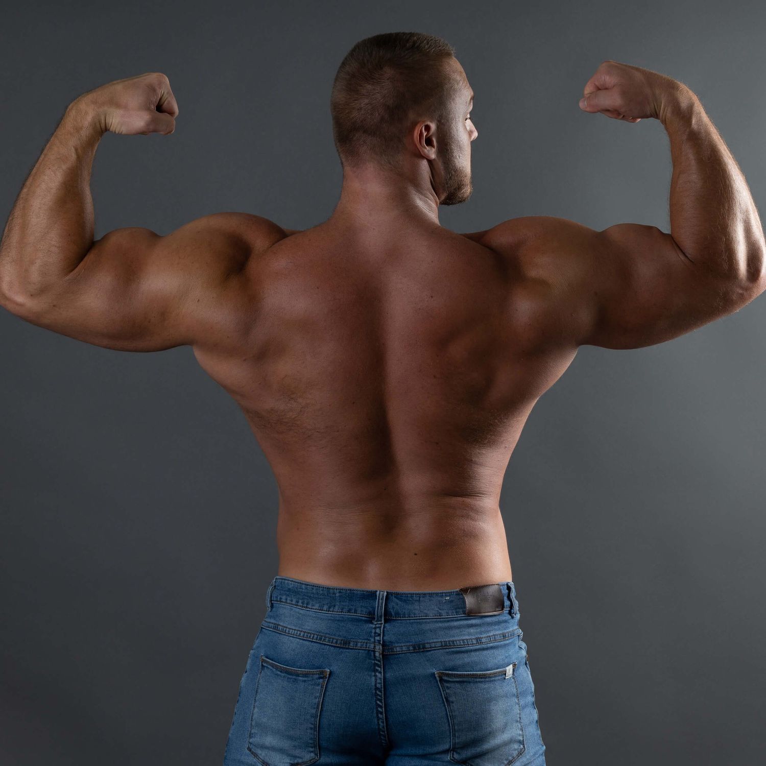 Muscular man flexing back muscles in blue jeans on dark background.