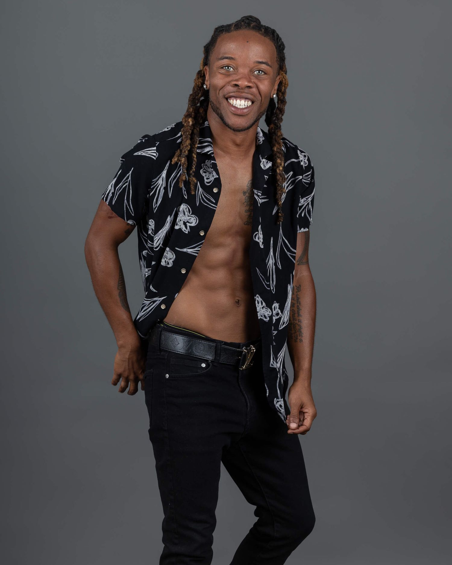 Smiling man with braids wearing open floral shirt and black jeans showing toned abs.