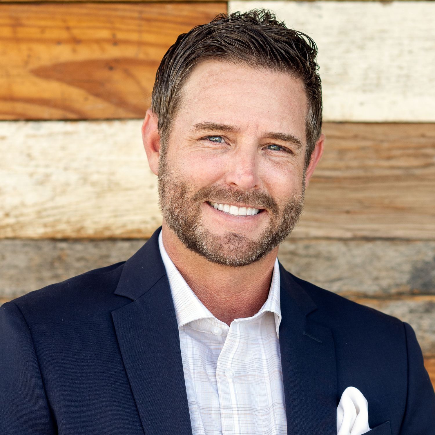 Smiling bearded man in navy blazer poses for professional headshot against rustic wood background.