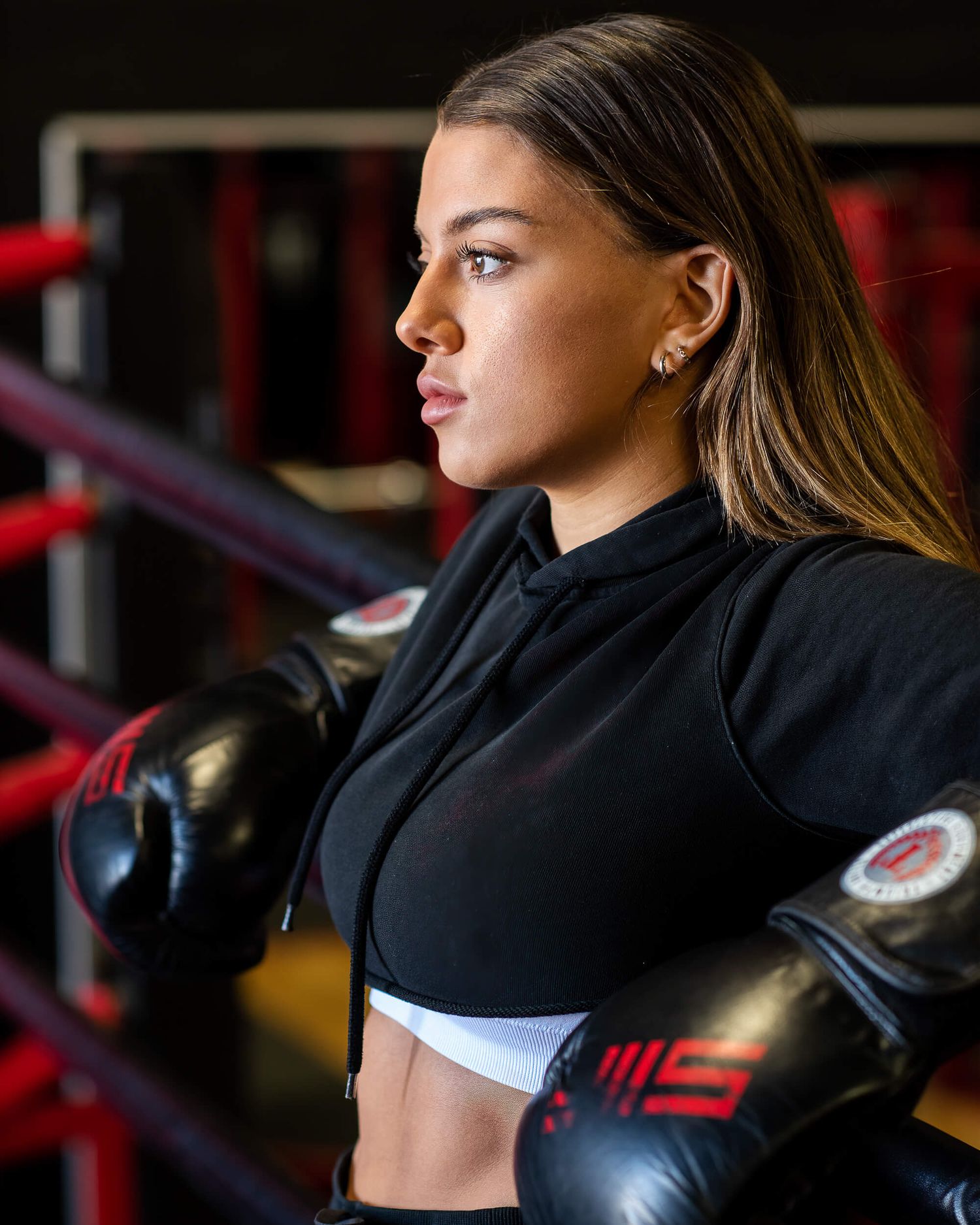 Female boxer in black hoodie and boxing gloves posing in a gym ring.