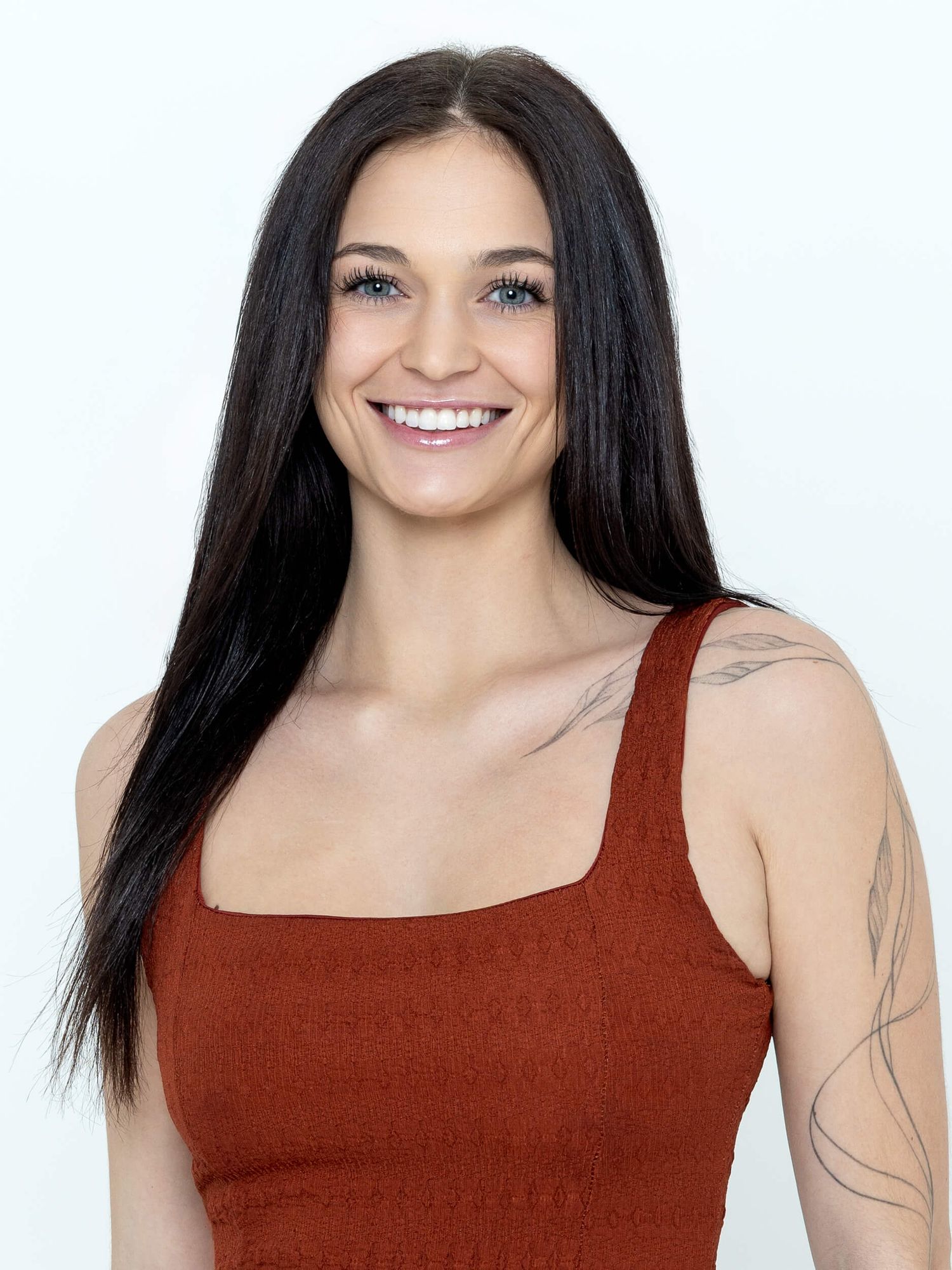 Smiling young woman with long dark hair wearing a rust-colored tank top, showcasing a floral arm tattoo.