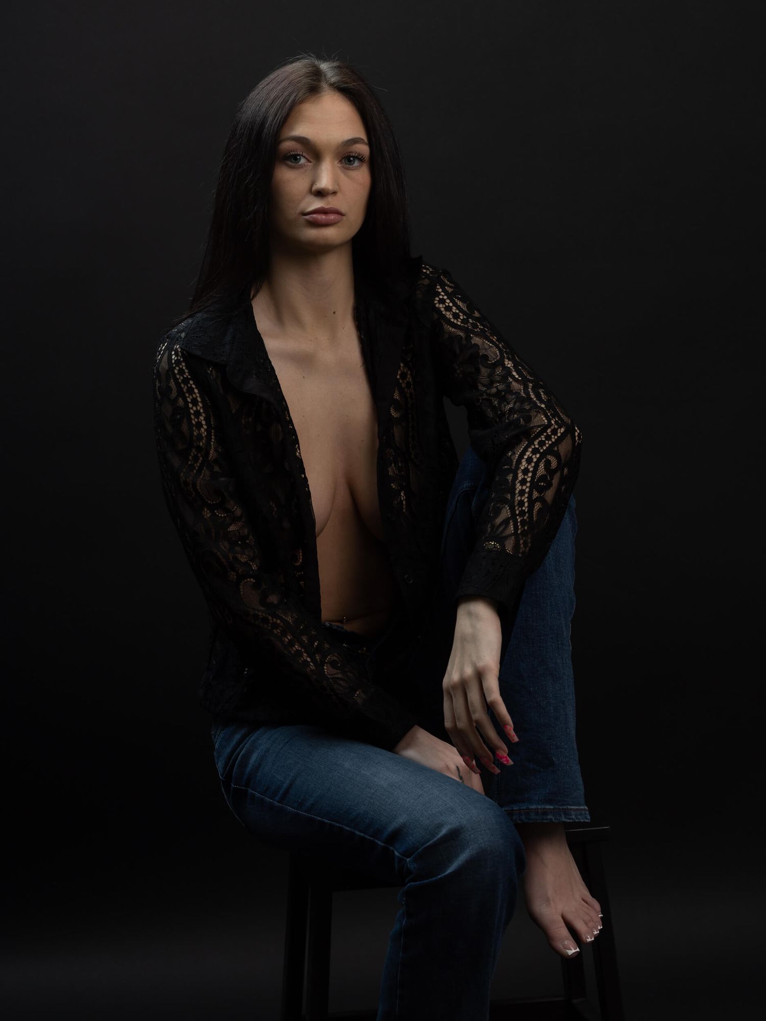 Woman in black lace jacket and jeans seated against dark background in a studio portrait.