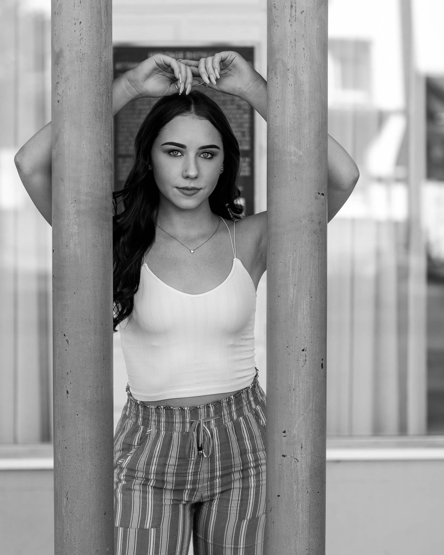 Young woman in white tank and striped pants posing between wooden pillars in black and white photo.