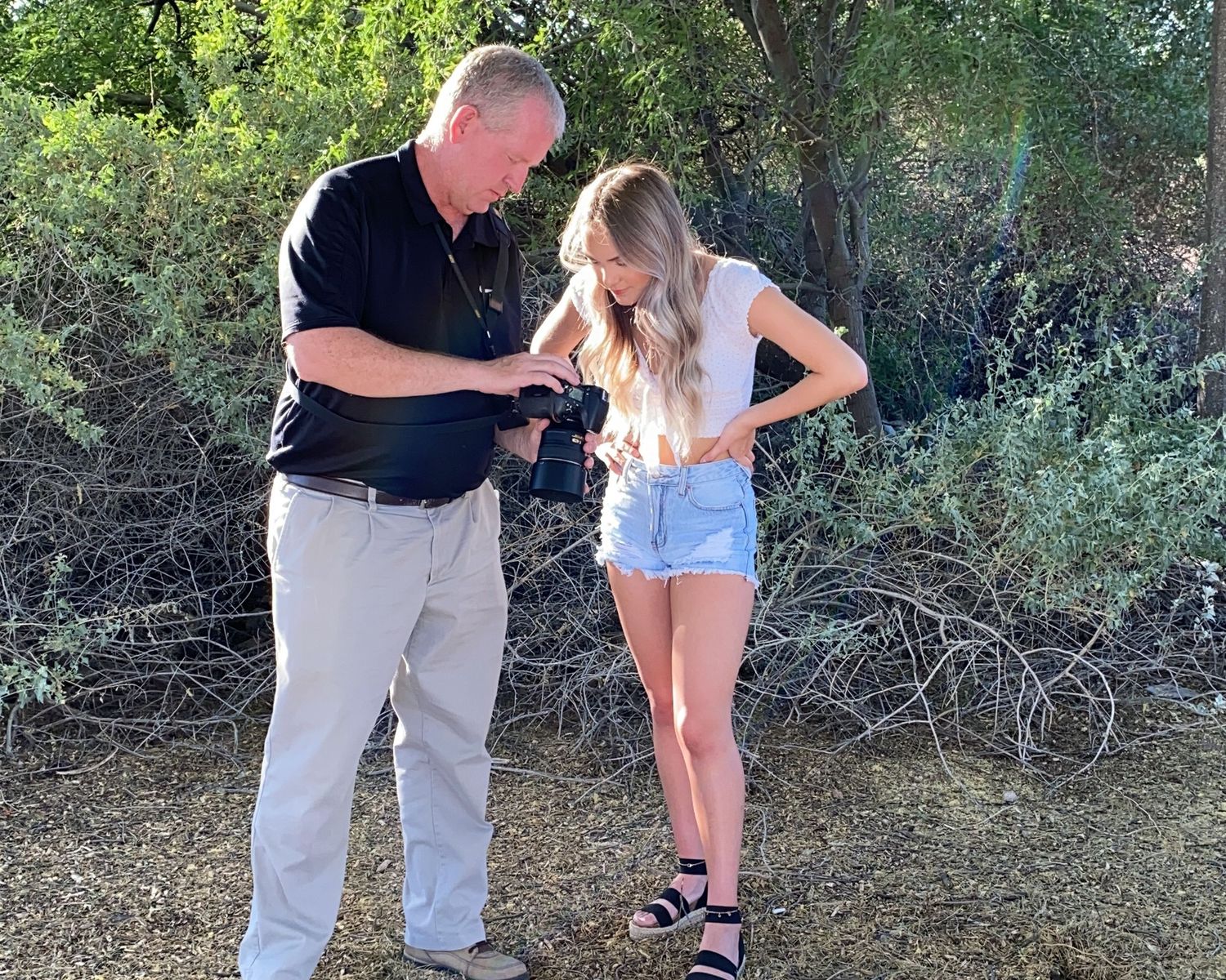 Photographer reviews camera with young blonde female model outdoors among green shrubs and trees.