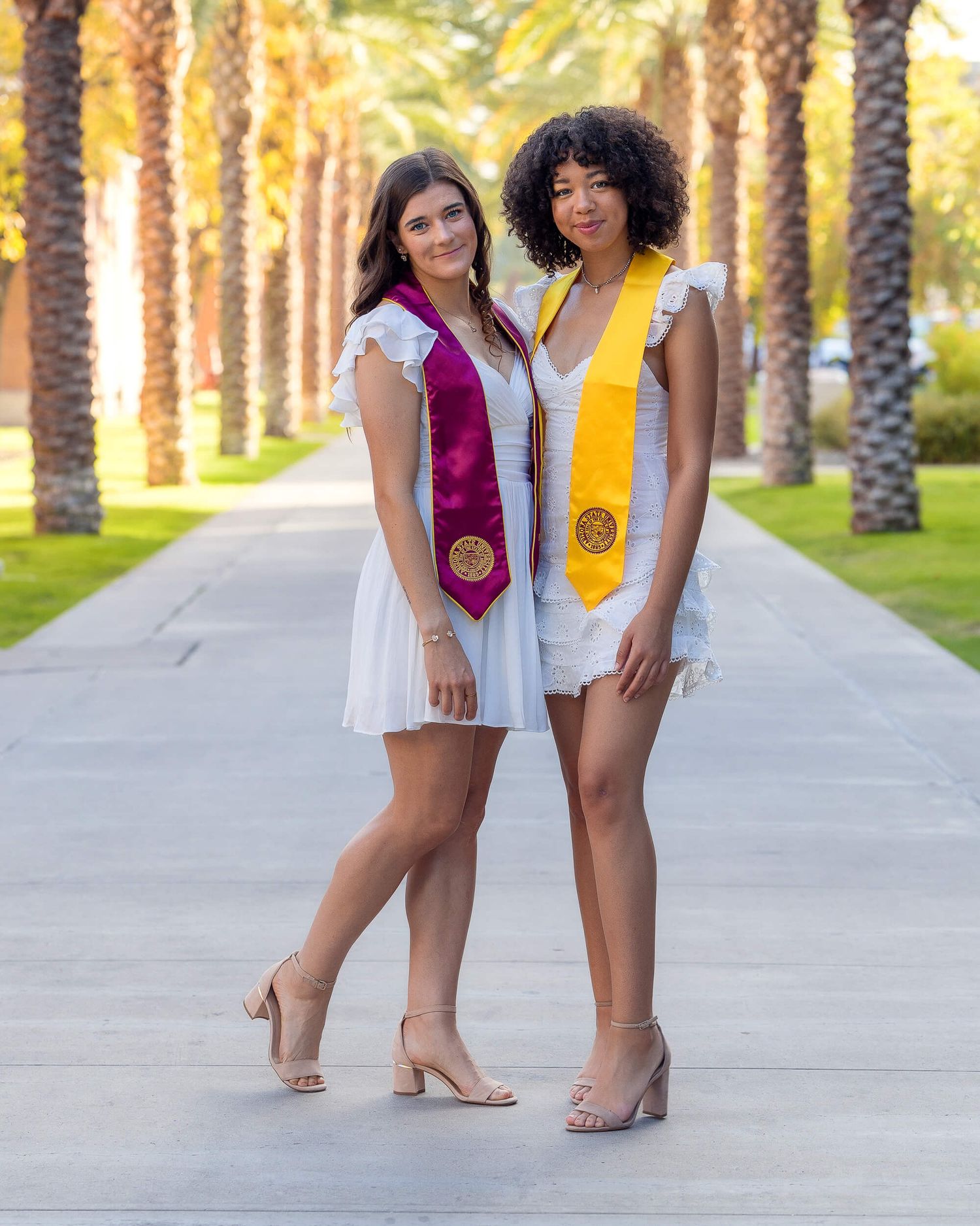 Two female graduates pose in white dresses with maroon and gold graduation stoles among palm trees.