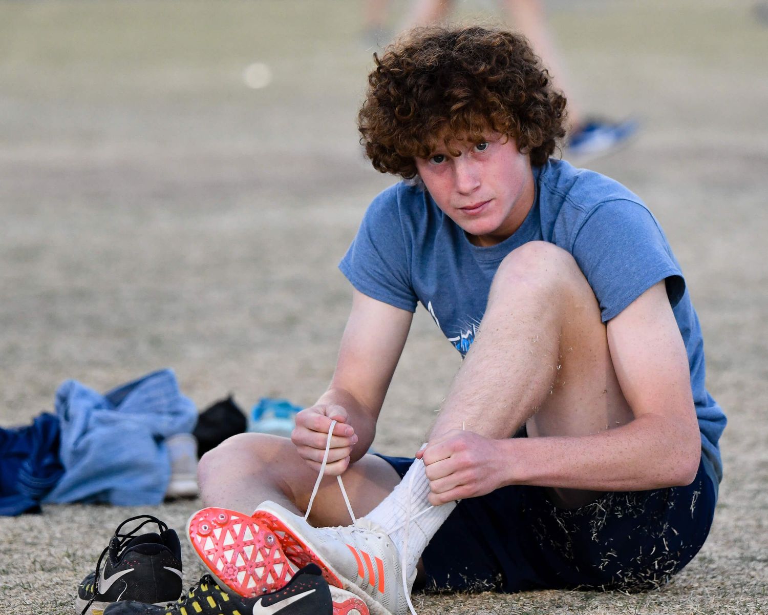 Curly-haired male athlete in blue shirt sits on the ground tying his track spikes before a competition.