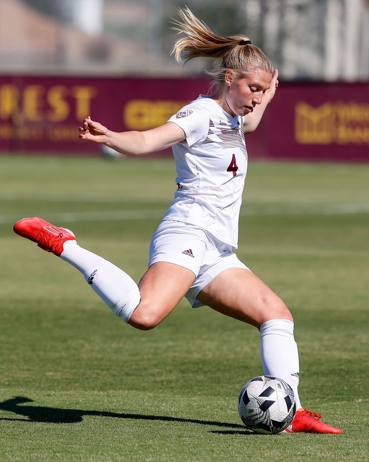 Female soccer player in white number 4 jersey prepares to kick the ball on a sunny field.