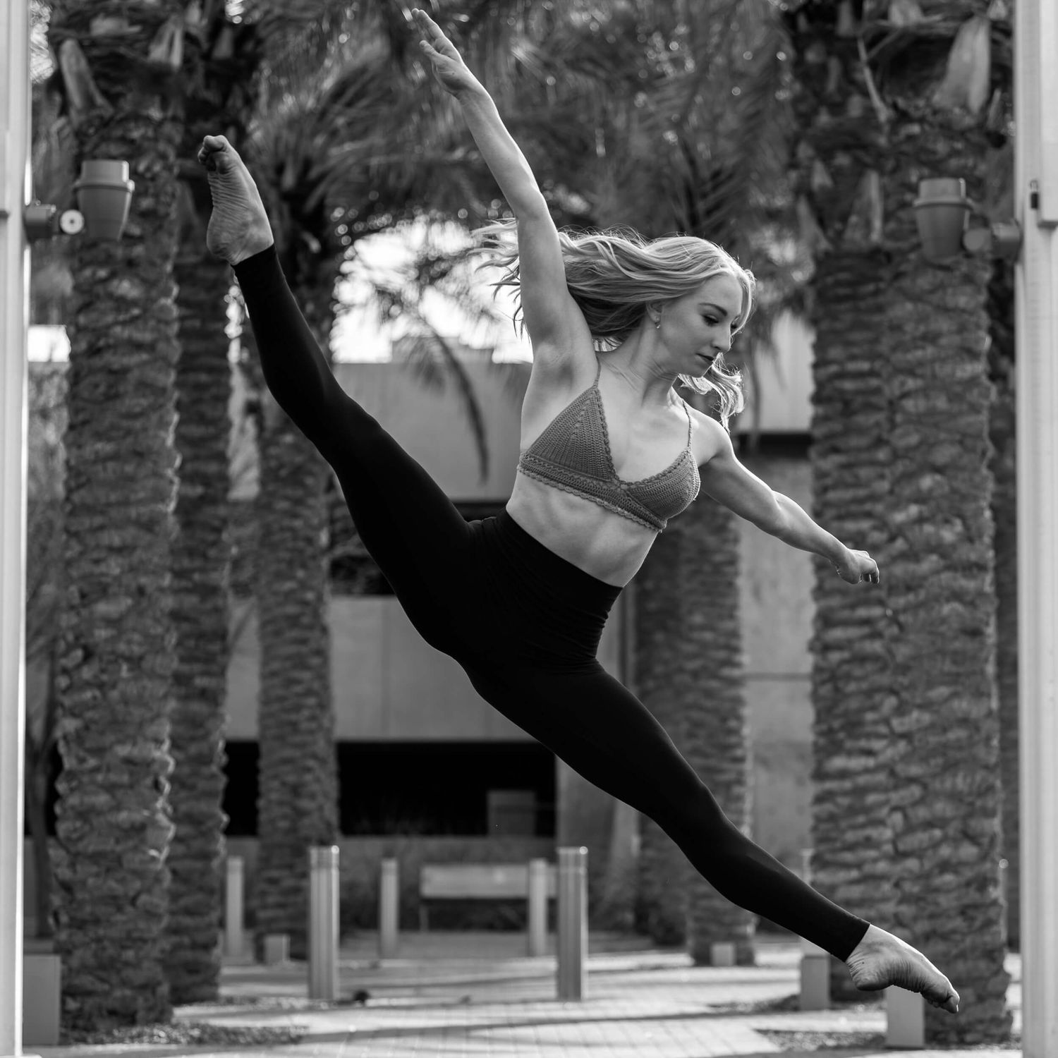 Blonde dancer performs aerial split leap outdoors near palm trees in black and white photo.