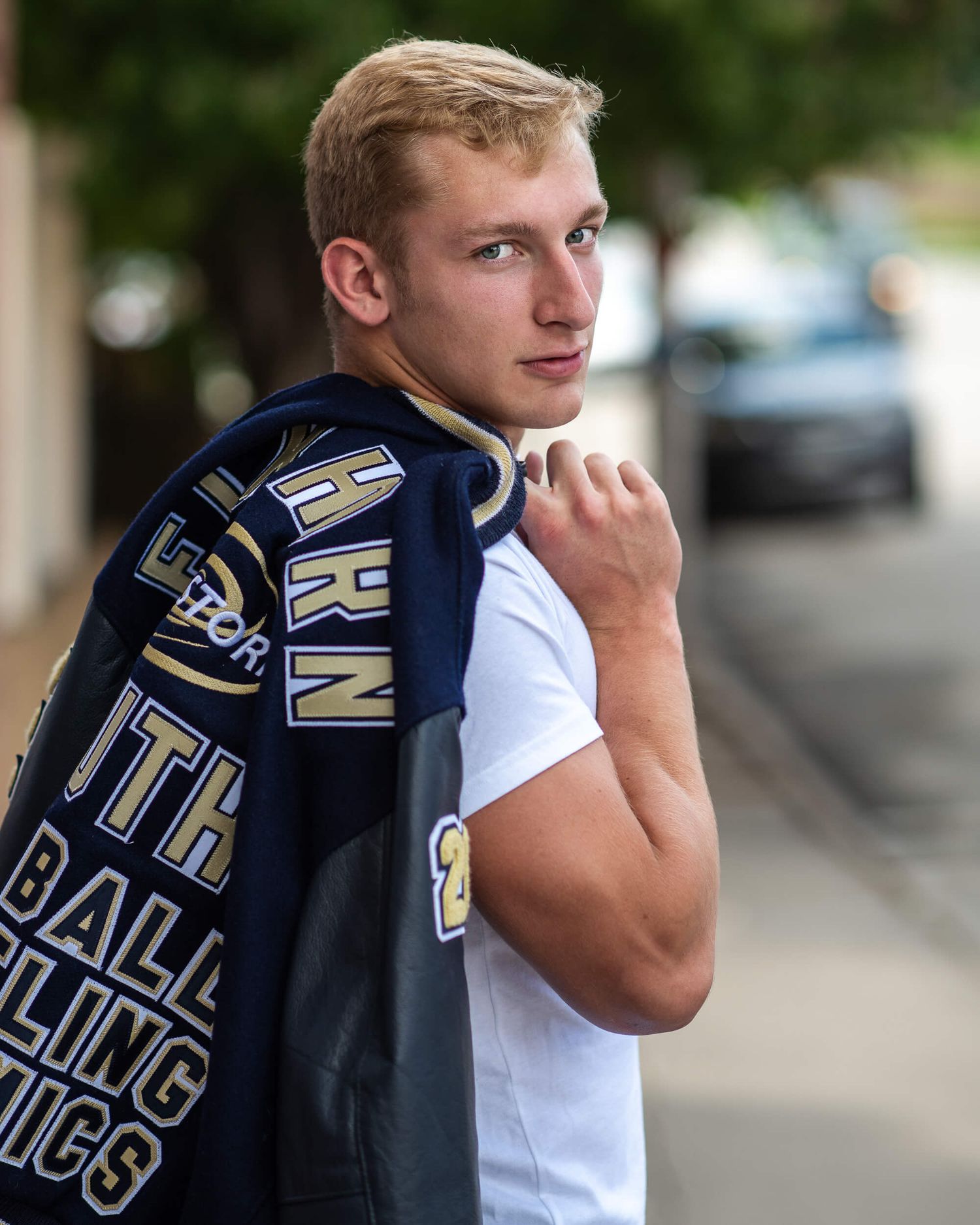 Blonde teen male senior photo wearing letterman jacket over shoulder on urban street.