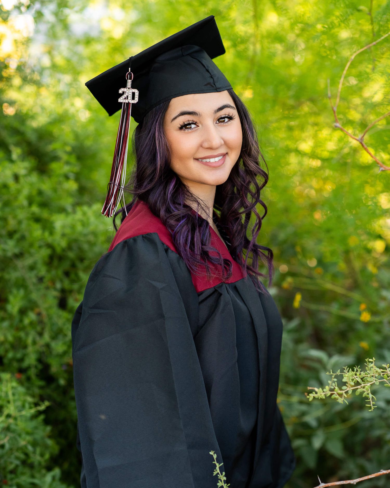 A smiling graduate in black cap and gown with red collar poses outdoors among lush green foliage.