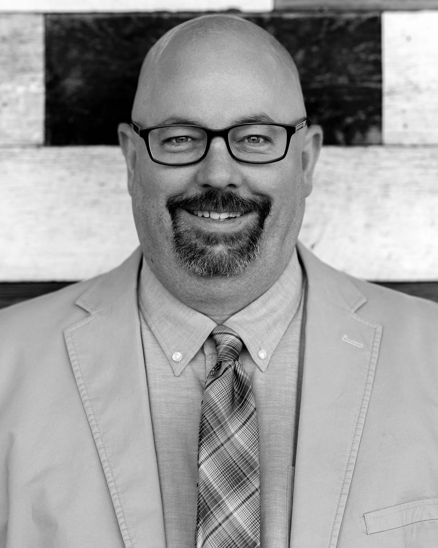 Bald man with glasses and beard wearing a blazer and plaid tie, smiling in a professional headshot photo.