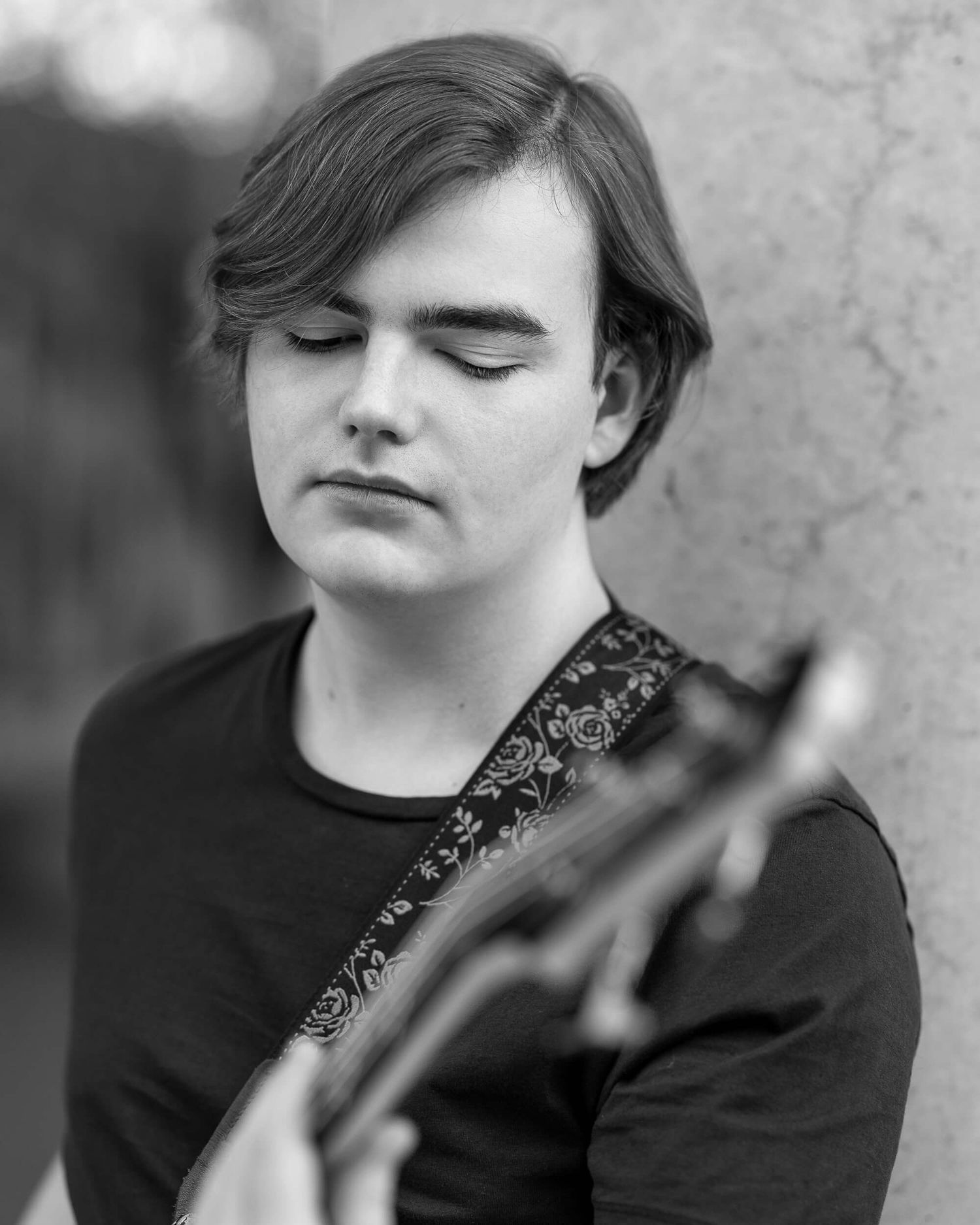 Black and white portrait of young musician with eyes closed holding a guitar outdoors.