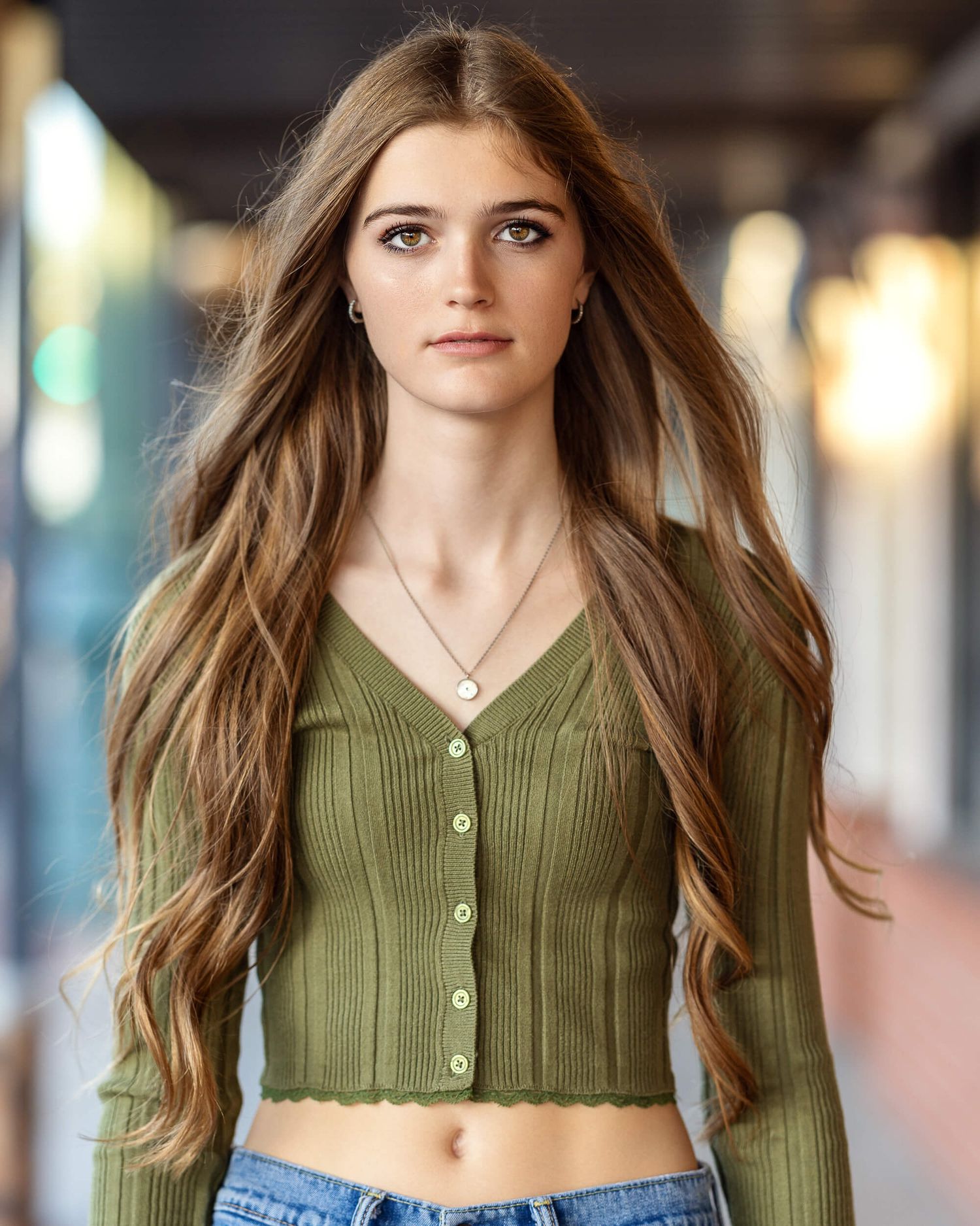 Young woman with long wavy brown hair wearing olive green cardigan crop top and jeans.